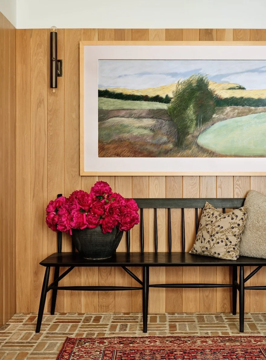 Entryway in a renovated home in Dorset, Vermont, with vertical wood wall paneling, a wooden bench, framed landscape artwork, and a vase of pink flowers.