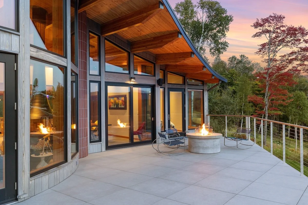 Terrace outside a custom home in Wilmington, Vermont, with a curved roofline, floor-to-ceiling glass, and a fire feature overlooking the landscape at dusk.