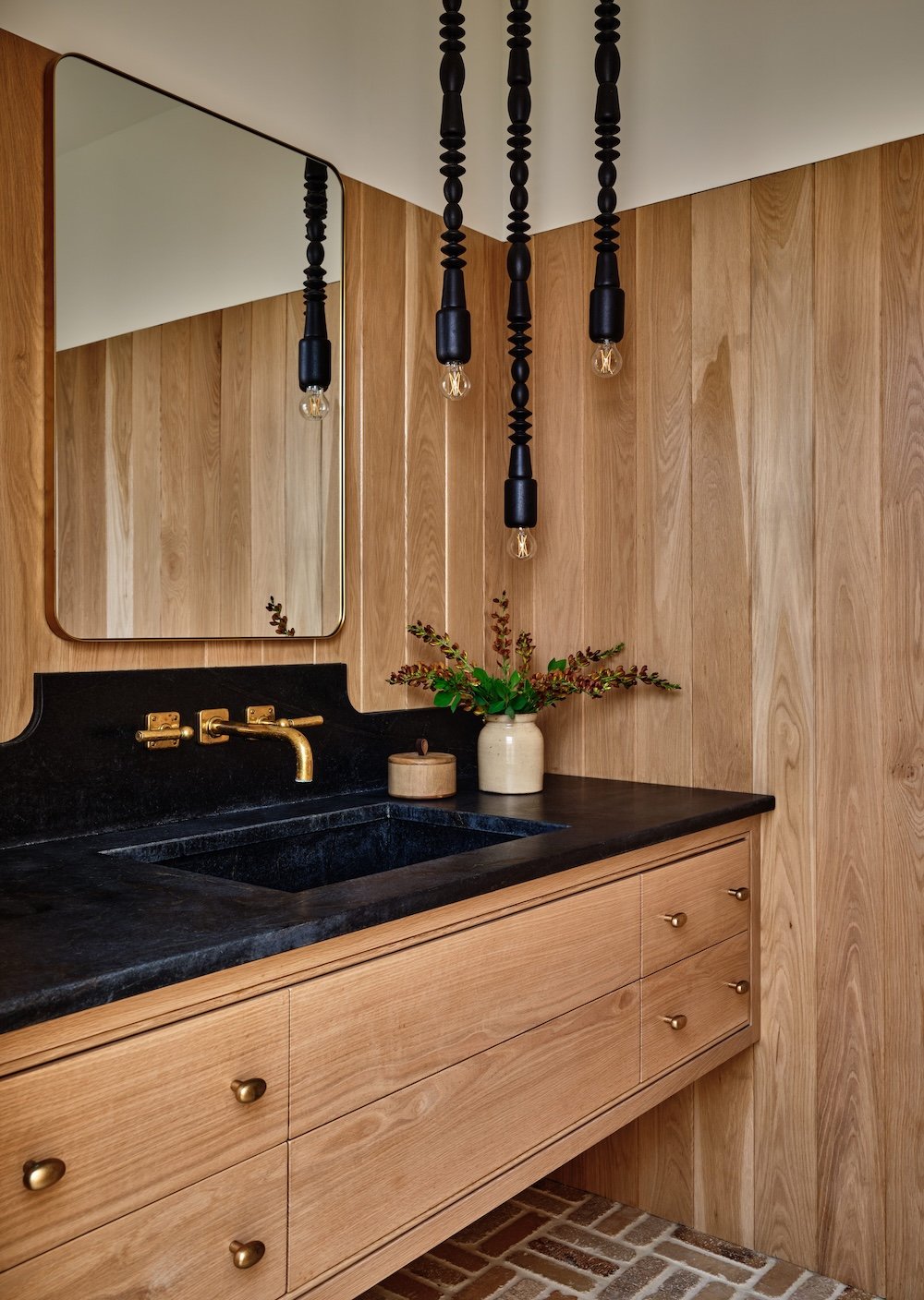 Bathroom vanity in a renovated home in Dorset, Vermont, with light wood cabinetry, paneled walls, a black countertop, and hanging pendant lights.