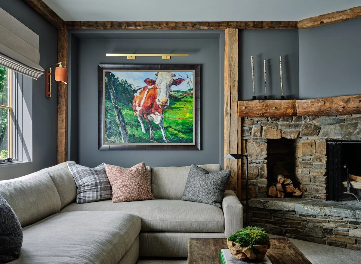 Living room in a renovated home in Dorset, Vermont, with a stone fireplace, exposed wood beams, built-in shelving, and a sectional beneath a large framed painting.