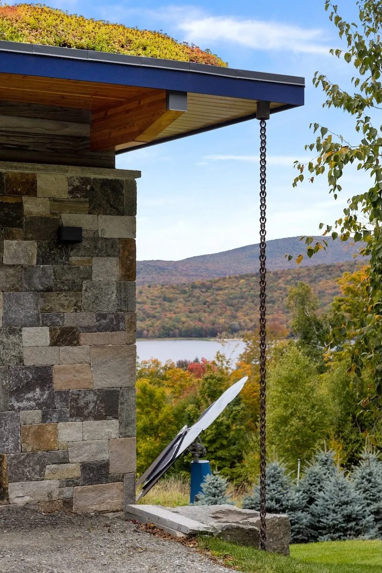 Side exterior of a custom home in Wilmington, Vermont, with stone facade, deep roof overhangs, and views toward the lake.