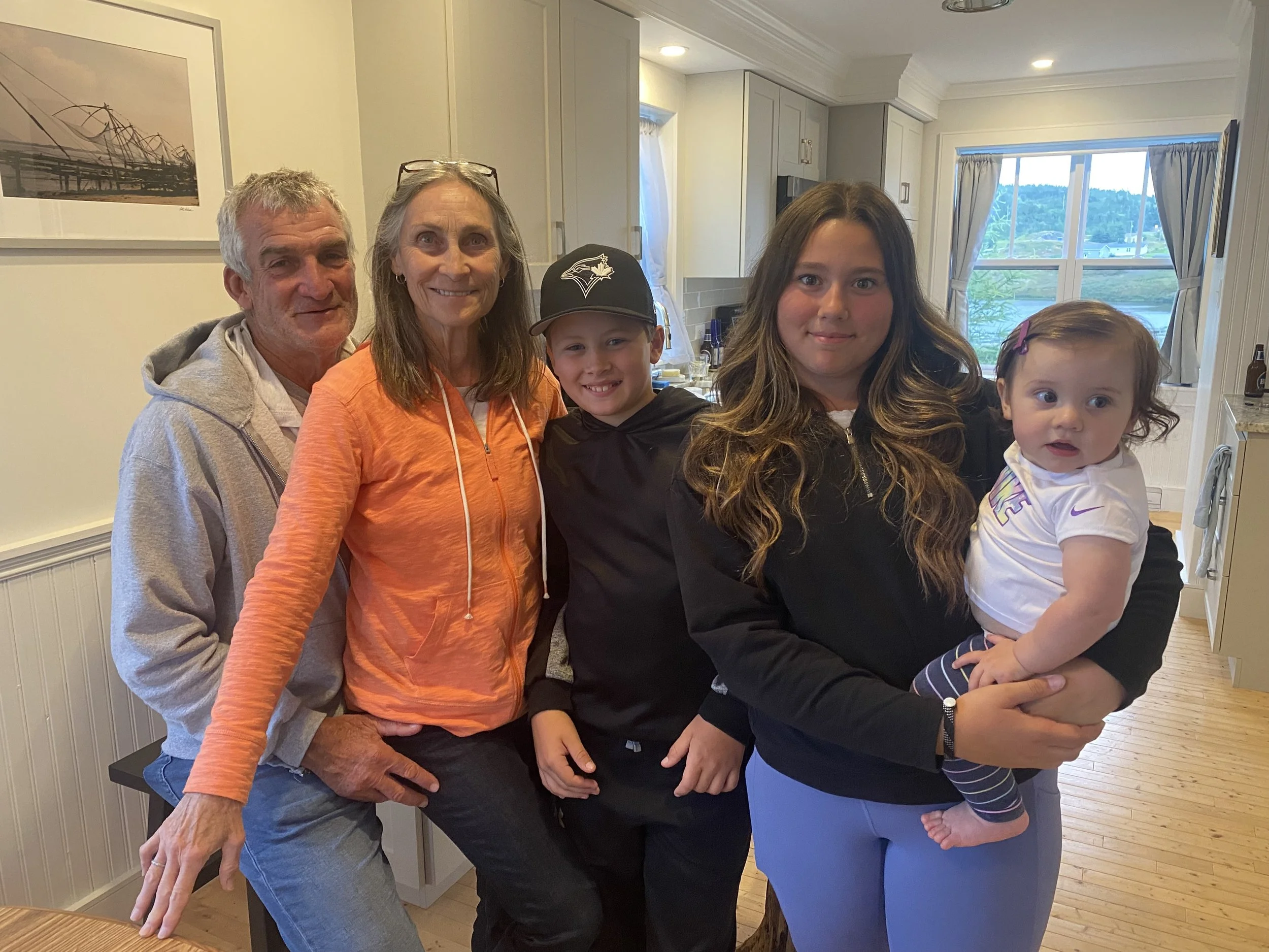 A multigenerational family group photo in a kitchen with five people: an older man, an older woman, a young girl, a teenage girl, and a toddler girl, standing close together and smiling.