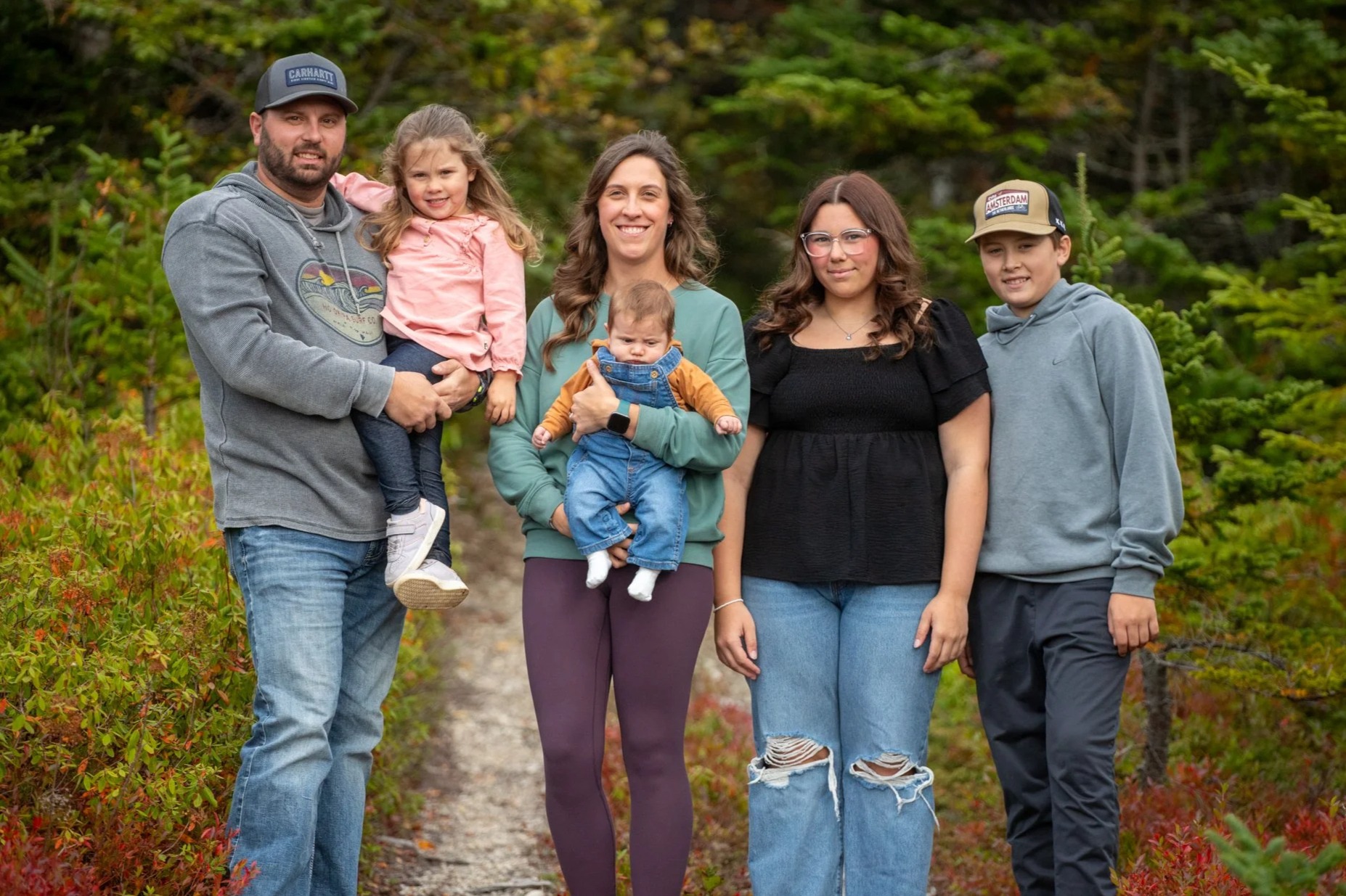 A family walking outdoors on a trail surrounded by green and red autumn foliage. The group includes two adults, two teenagers, and two young children, all smiling and dressed casually.