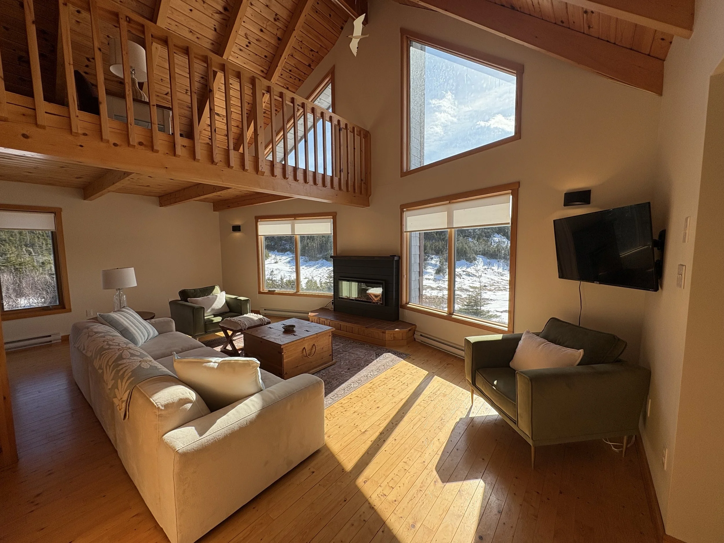 Living room with large windows, high wooden ceiling, wooden floors, and furniture including a beige sofa, green armchair, wooden coffee table, fireplace, and wall-mounted flat-screen TV.
