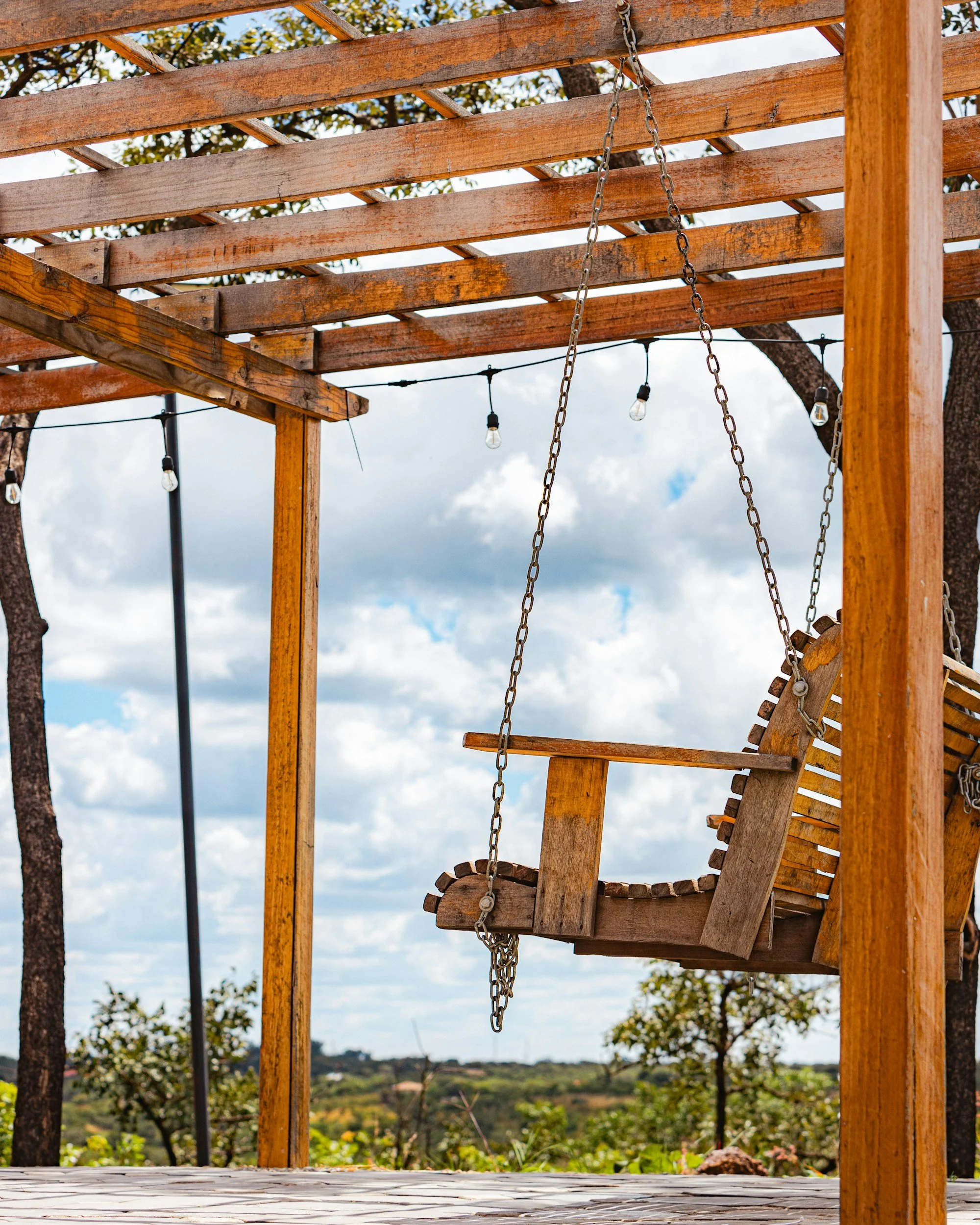 A wooden structure with swings hanging from chains outdoors, with trees and a partly cloudy sky in the background.