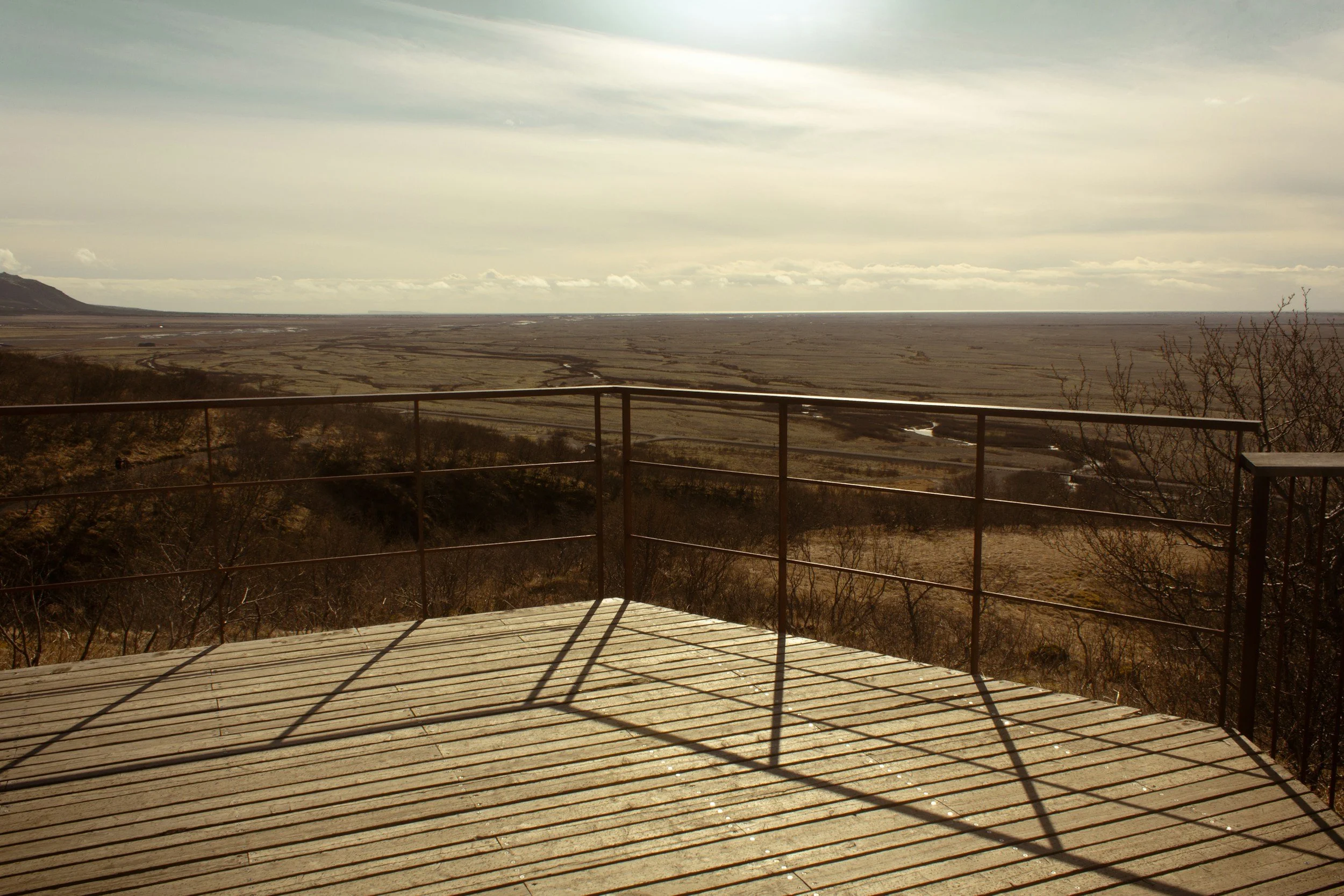 A wooden deck with a metal railing overlooking a vast, flat coastal landscape under an overcast sky. Sparse leafless trees are visible at the edge of the deck.