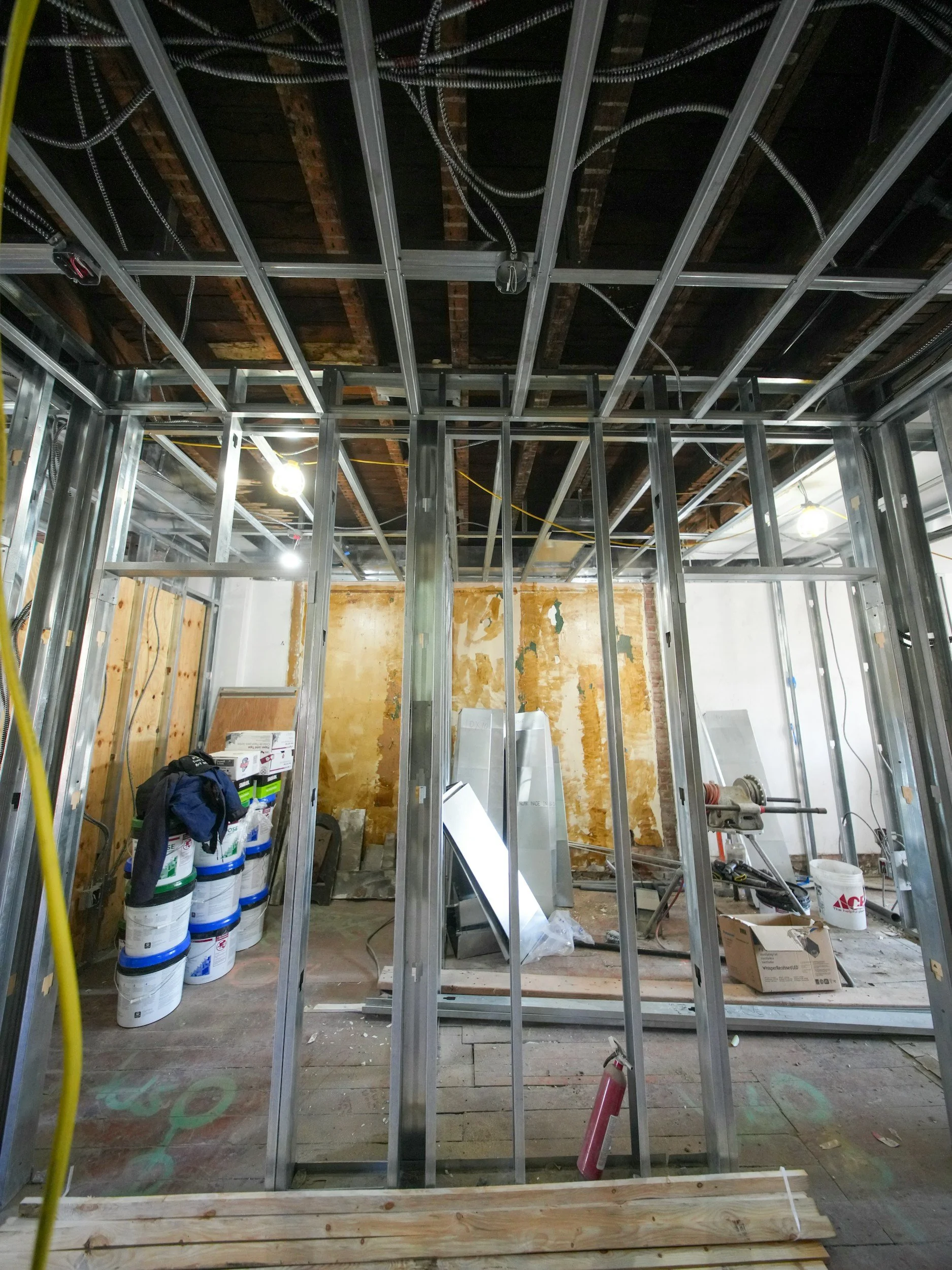 Interior view of a building under construction, showing metal framing, electrical wiring, and construction tools and materials.