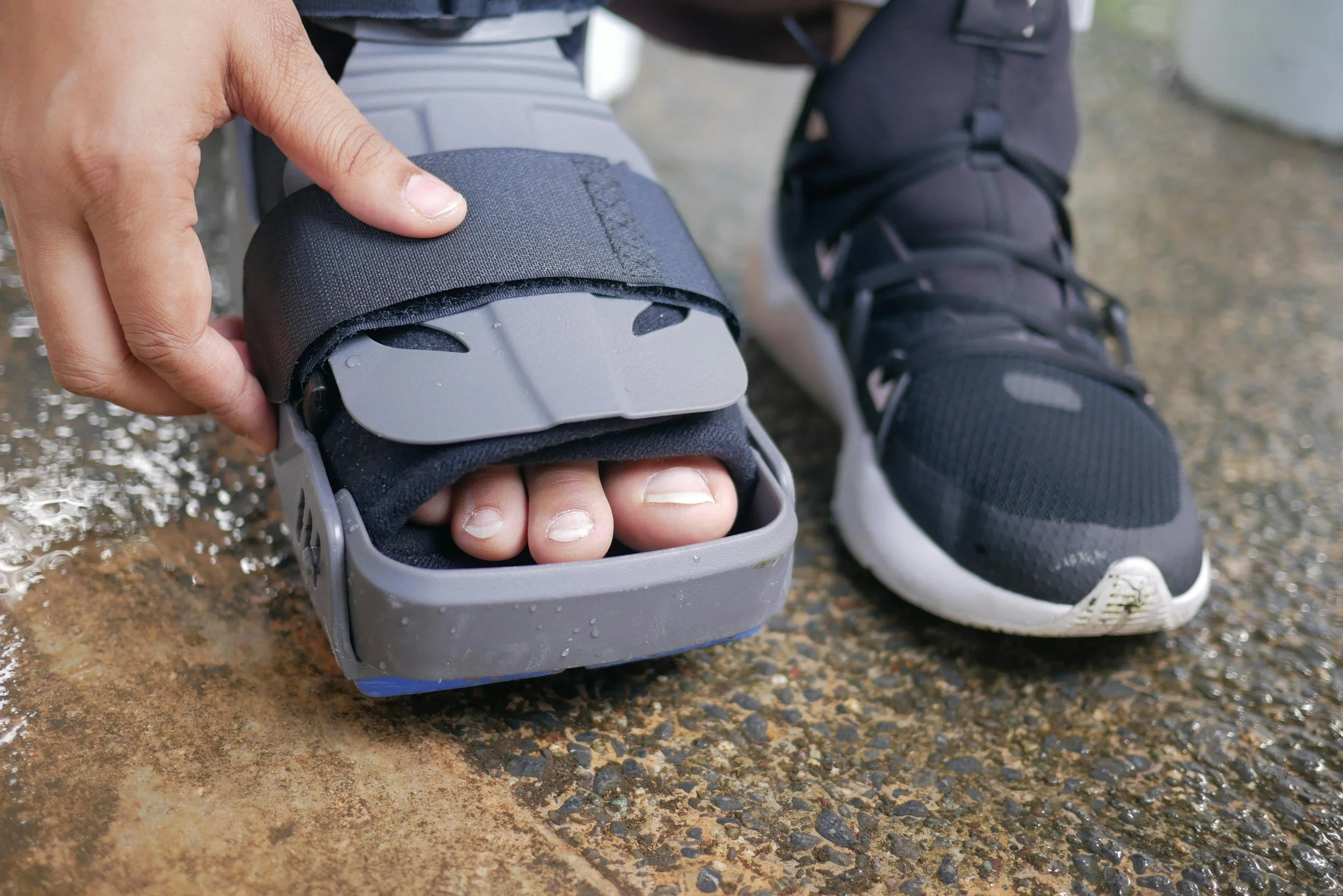 Someone adjusting a roller skate on their foot, with black athletic shoes and laces, on a wet, rough pavement surface.