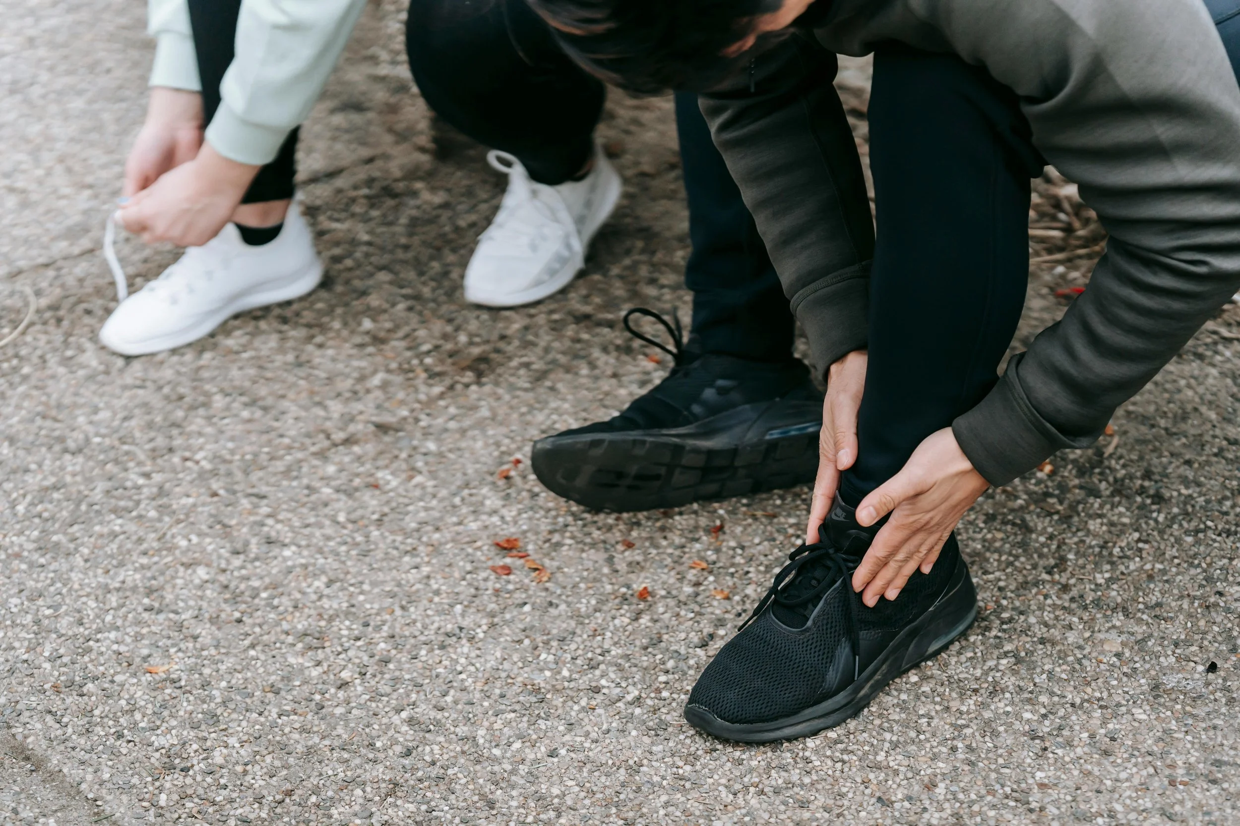 Two people are tying their shoelaces on a paved surface, one wearing black running shoes and the other wearing white sneakers.