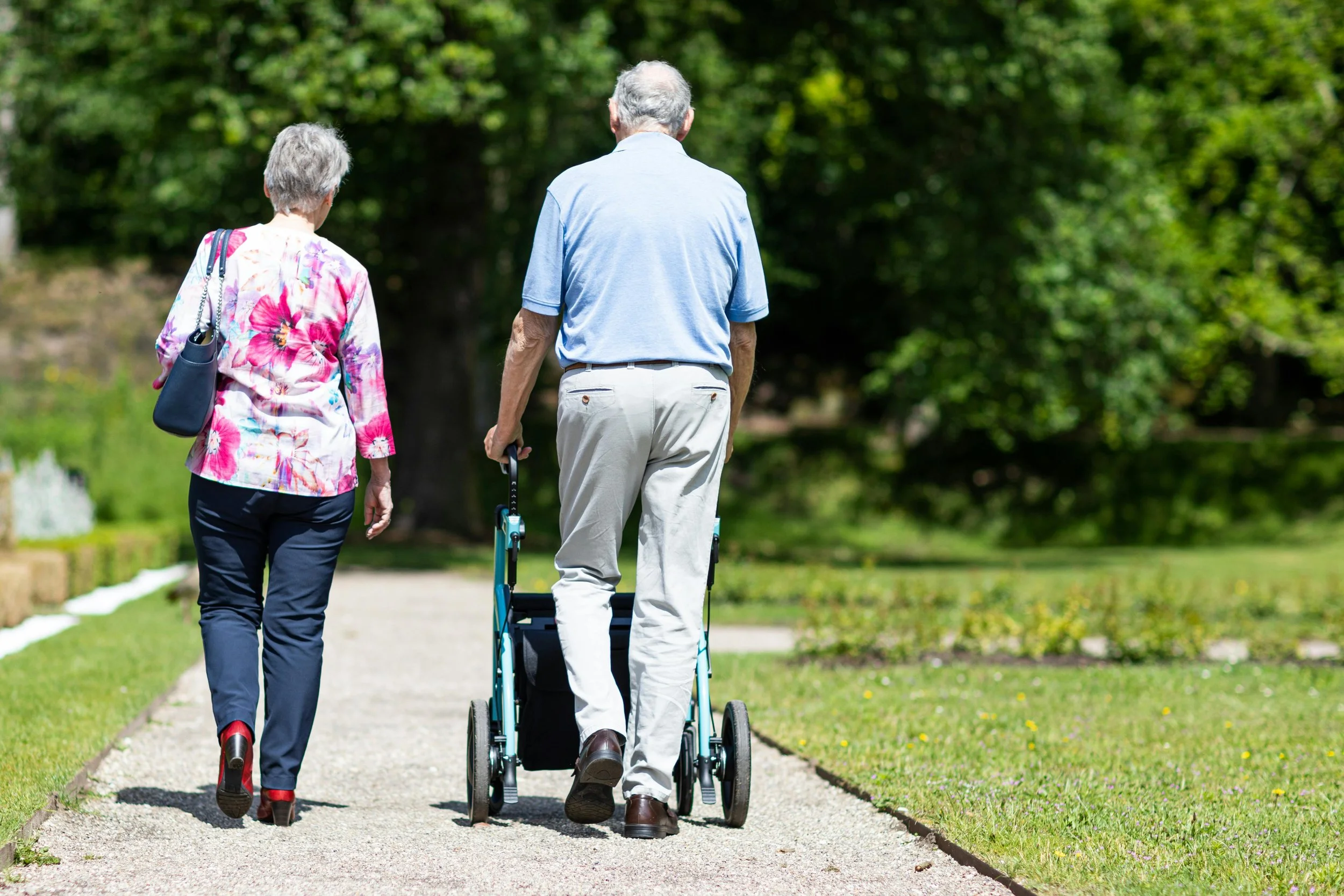 An elderly man in a blue polo shirt and beige pants walking with a cane, accompanied by an elderly woman in a floral blouse and dark pants, walking together on a paved path in a park with green trees in the background.