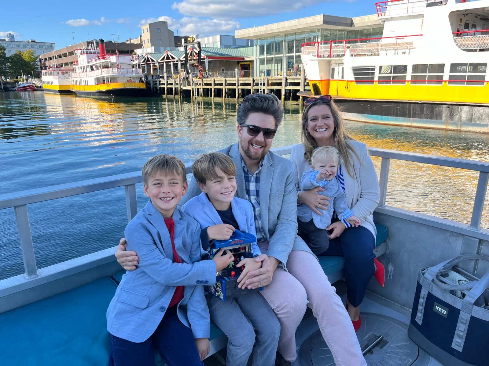 A family of five sitting on a boat near a dock with boats and buildings in the background. They are smiling and enjoying a day out.