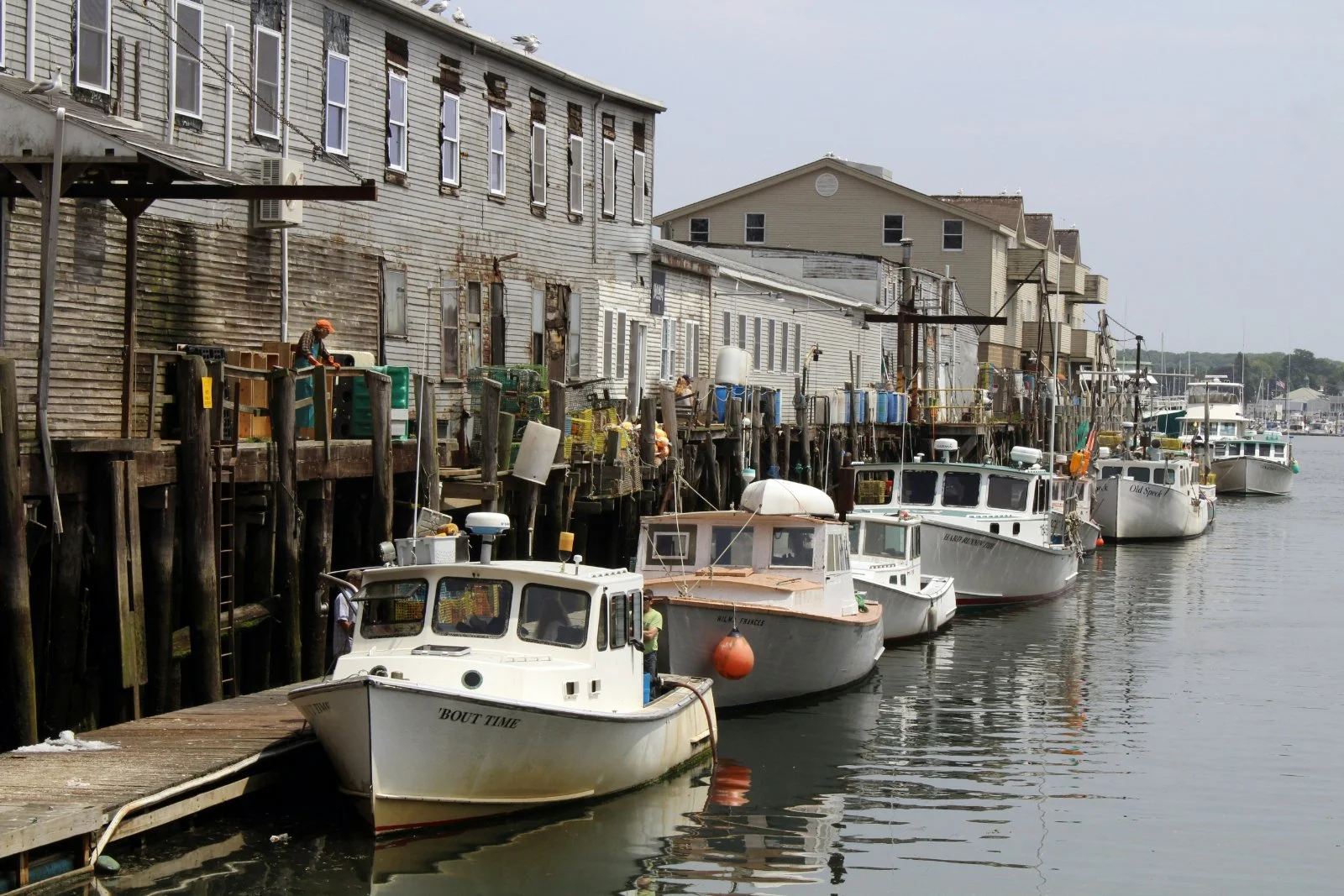 Boats docked along a weathered wooden pier near old, gray buildings lining a harbor.