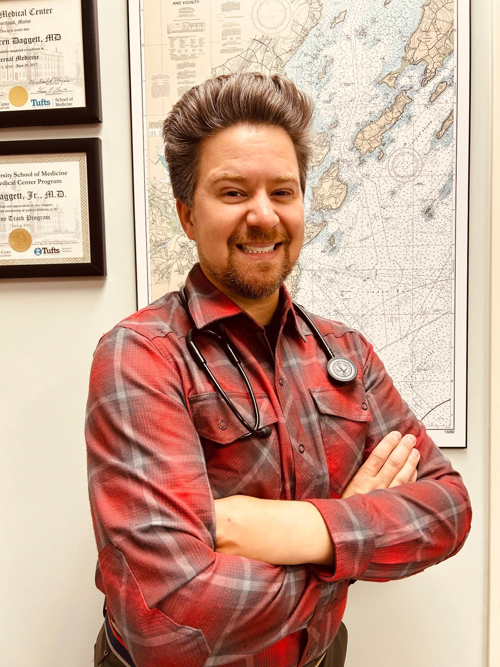 A man with short brown hair and a beard, smiling and crossing his arms, wearing a red plaid shirt and a stethoscope on his neck, standing in front of a medical map and framed certificates on the wall.