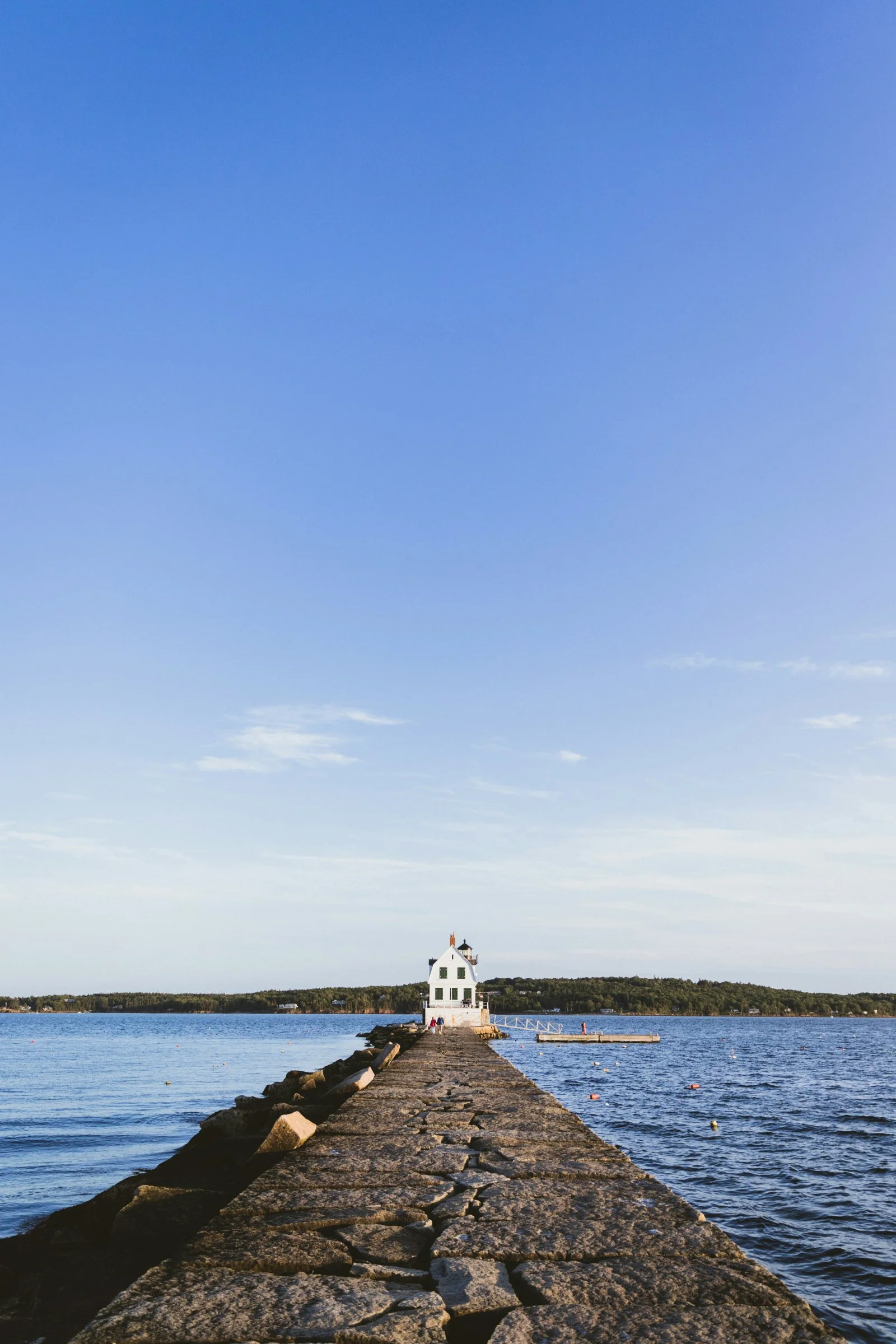A rocky pier extends into a body of water leading to a small white house with a pointed roof and chimney, situated on a strip of land with trees, under a clear blue sky.