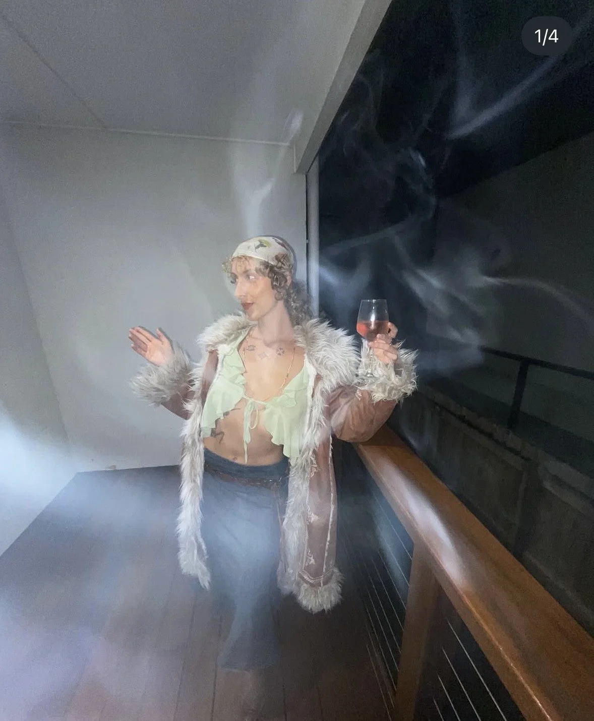 A woman dressed in a vintage 70s style with a sheer green top, fluffy coat, and headscarf, holding a glass of rosé wine, standing in a dimly lit room with smoky haze.