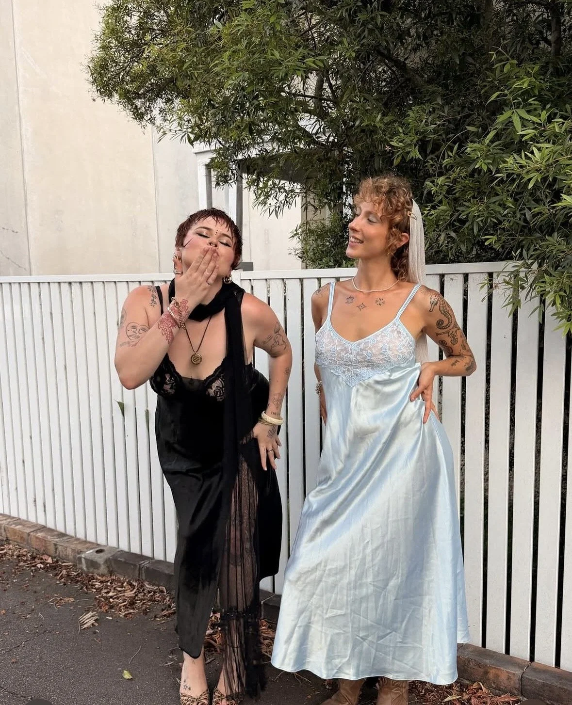Two women dressed in different styles standing outdoors near a white fence with trees in the background. One woman in black is blowing a kiss, and the other in a light blue dress is smiling.