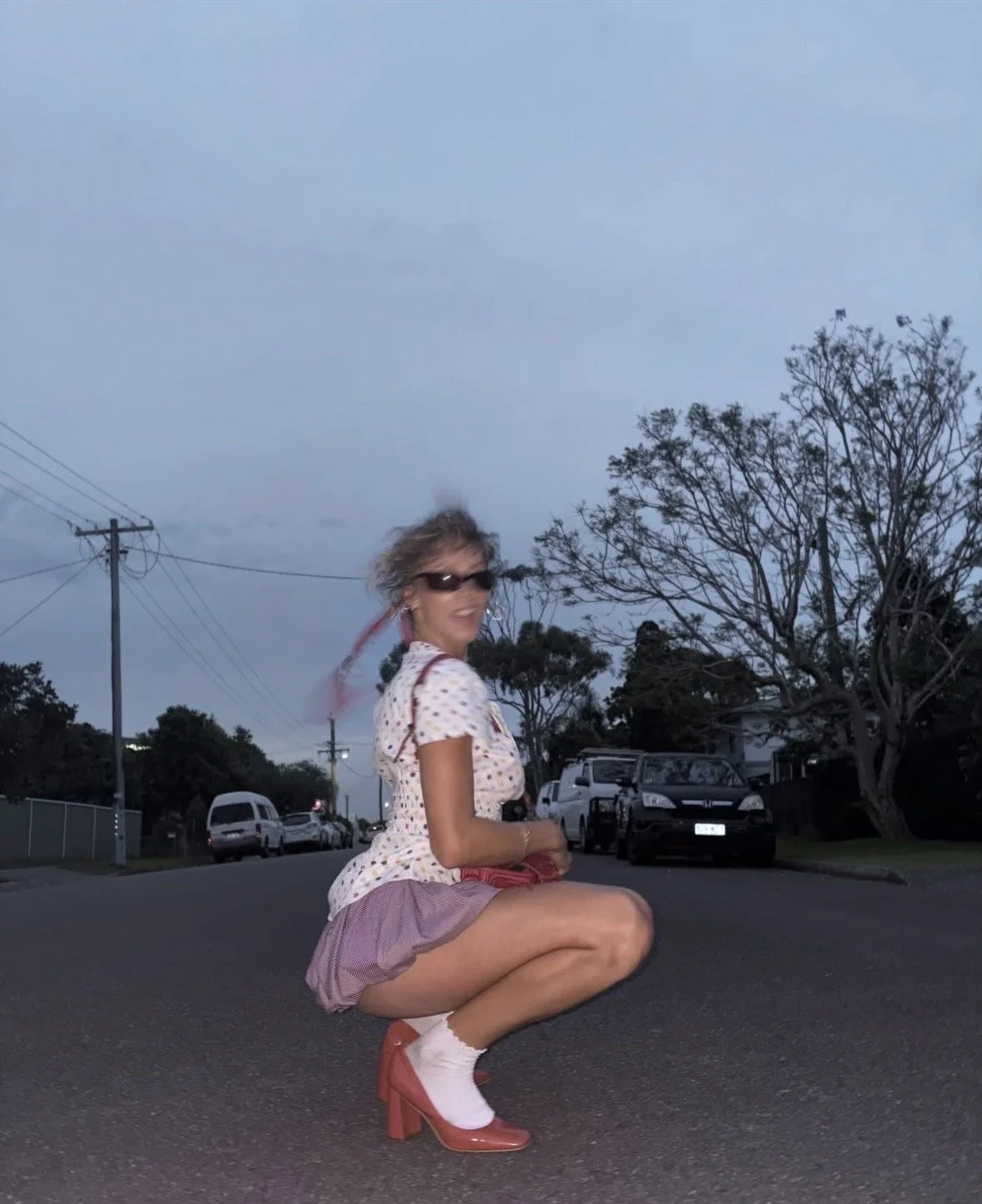 A woman squatting in the middle of a street during dusk, smiling at the camera. She is wearing sunglasses, a white polka-dot shirt, pink skirt, white socks, and red high heels. She has curly blonde hair and appears to be in a suburban area with parke