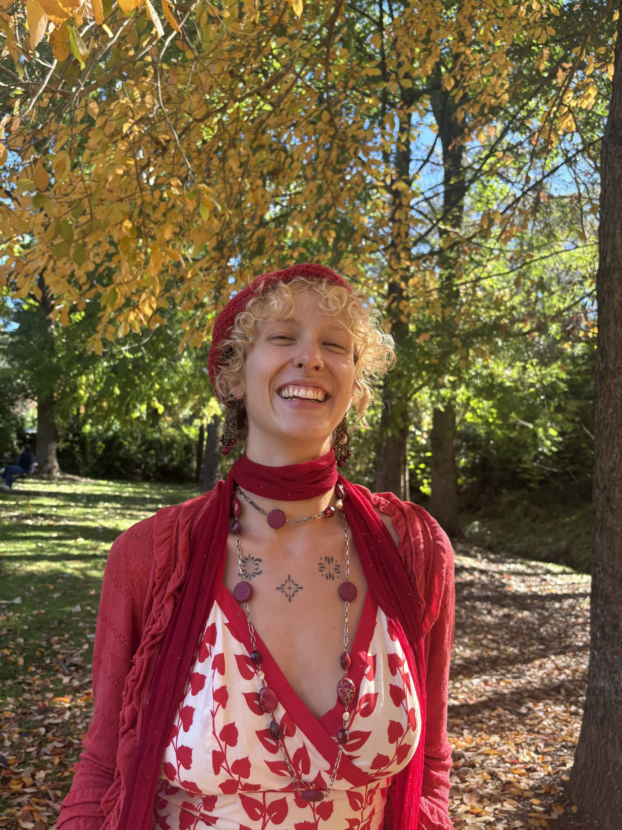 A smiling person with curly blond hair, wearing a red beanie, red cardigan, and a necklace with large beads, standing in a park with autumn trees and fallen leaves.