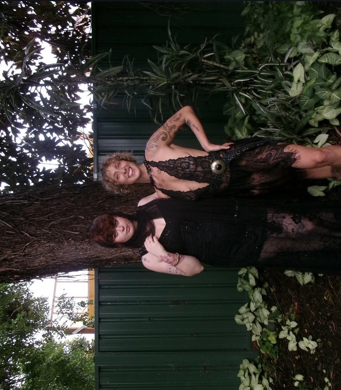 Two women standing outdoors in front of a green fence, surrounded by trees and plants. One woman is wearing a black lace dress with a large eye detail on the waist, smiling with curly hair. The other woman has dark short hair, tattoos on her arms, we