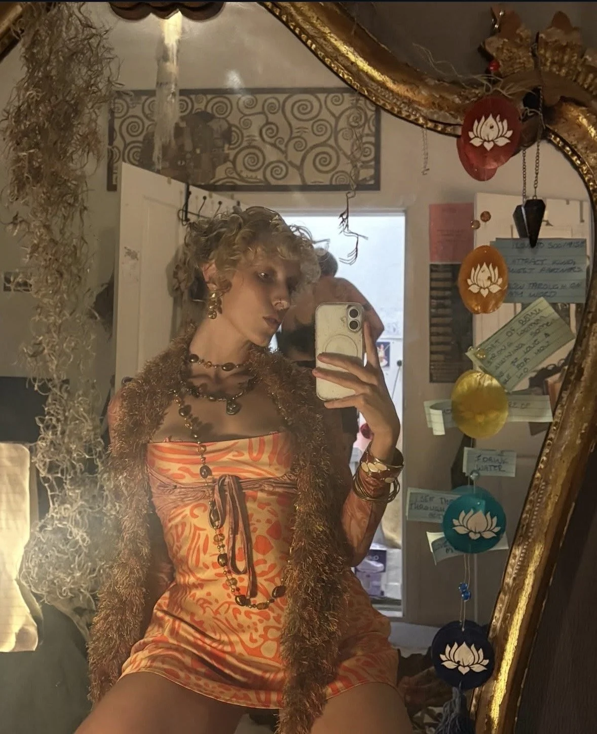 A woman taking a mirror selfie in a room decorated with Asian-inspired ornaments, including hanging lotus flower lanterns. She has curly hair and is wearing a colorful, patterned dress with layered necklaces and bracelets.