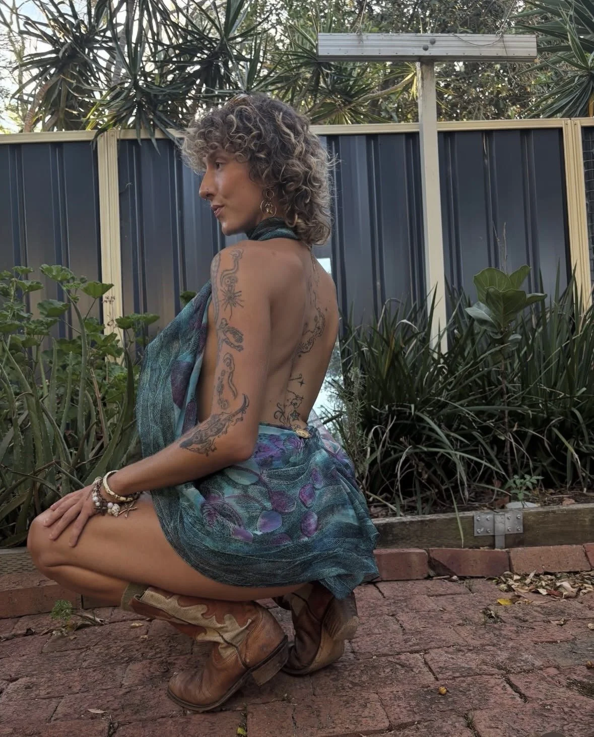 A woman with curly hair, tattoos on her arm and back, wearing a colorful wrap skirt and cowboy boots, squatting outdoors on a brick path in front of green plants and a metal fence.