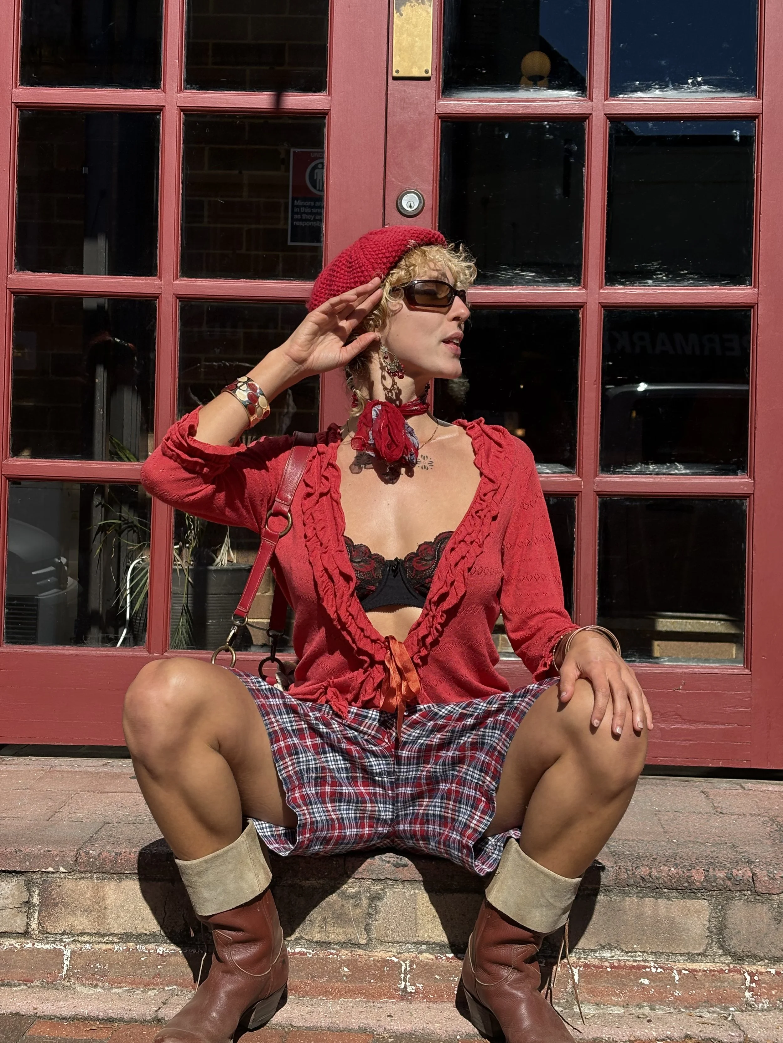 Person wearing a red beret, sunglasses, a red jacket, and plaid shorts, sitting on a brick sidewalk in front of a red door with glass panes, with their legs spread apart and one hand saluting.