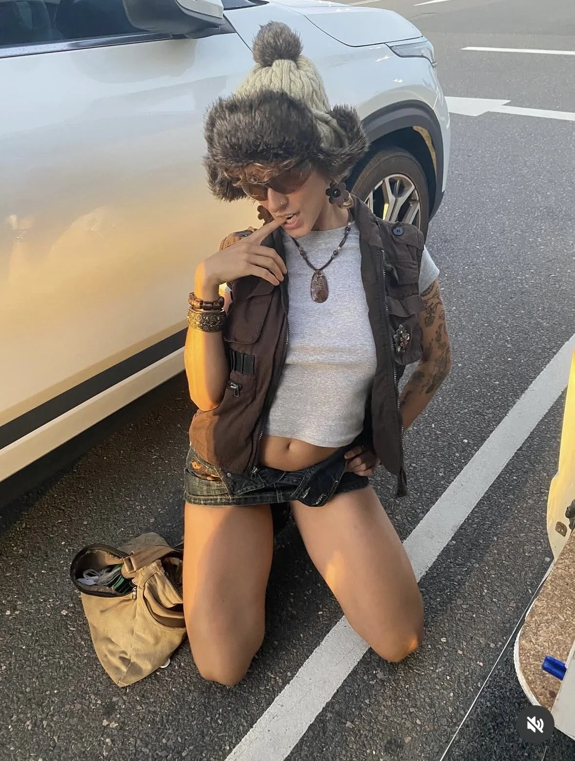A woman kneeling on the ground near a white vehicle, wearing a winter hat, sunglasses, a vest, a crop top, and shorts, touching her face with her right hand.