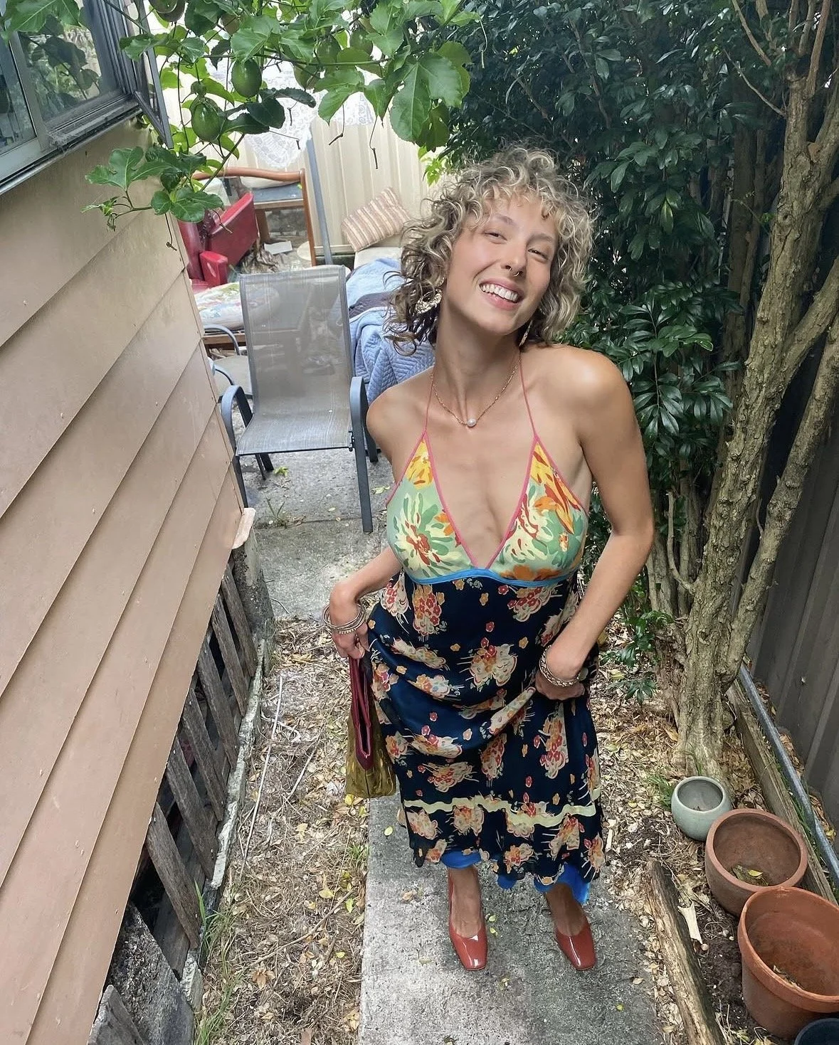 A smiling woman with curly blonde hair stands outdoors in a garden area, wearing a floral sundress and red shoes, with her hands in her pockets.