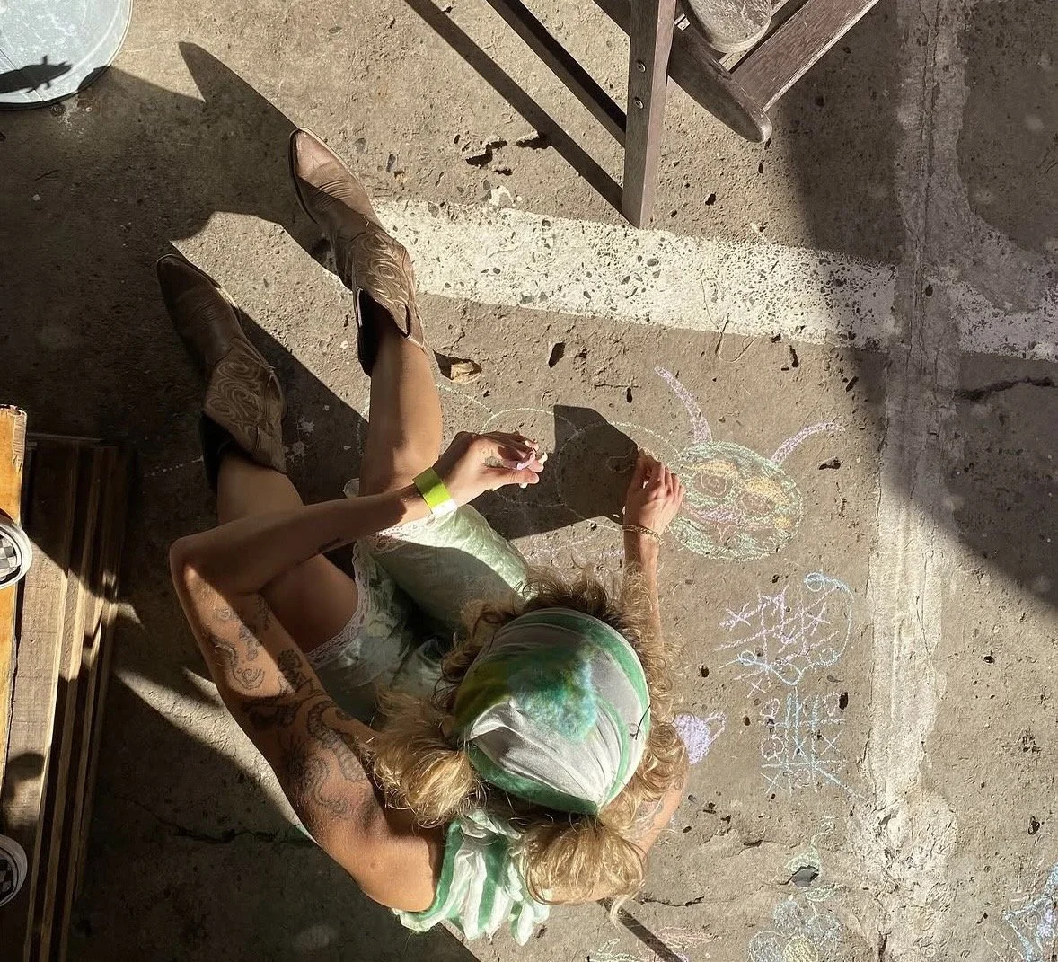 A person drawing a colorful chalk insect on a concrete floor, sitting with legs bent, wearing a tie-dye bandana, white dress, cowboy boots, and tattoos on arms. Sunlight creates shadows on the ground.