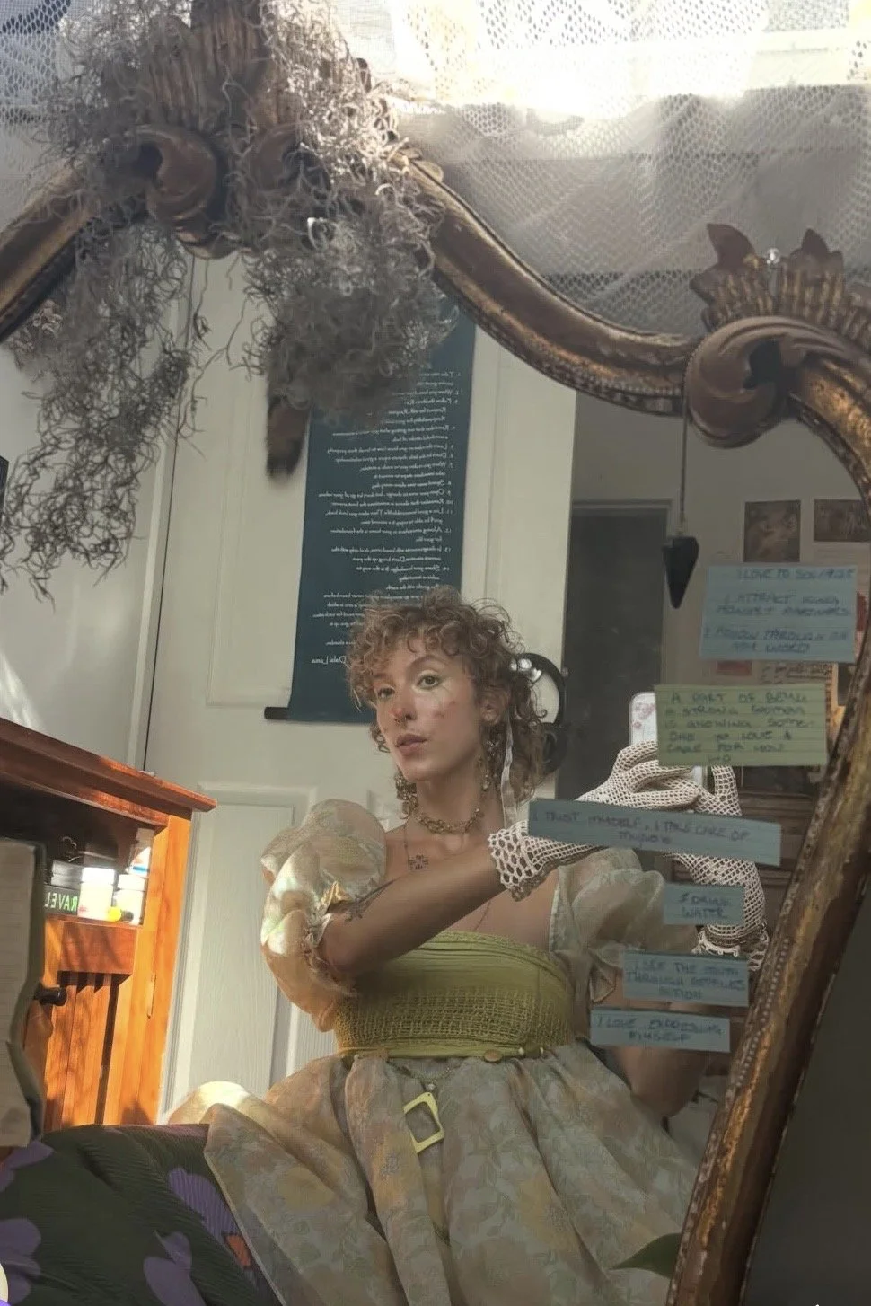 A woman with curly hair and vintage-style clothing taking a selfie in a mirror decorated with notes and ornaments, with a coat and other objects visible in the background.