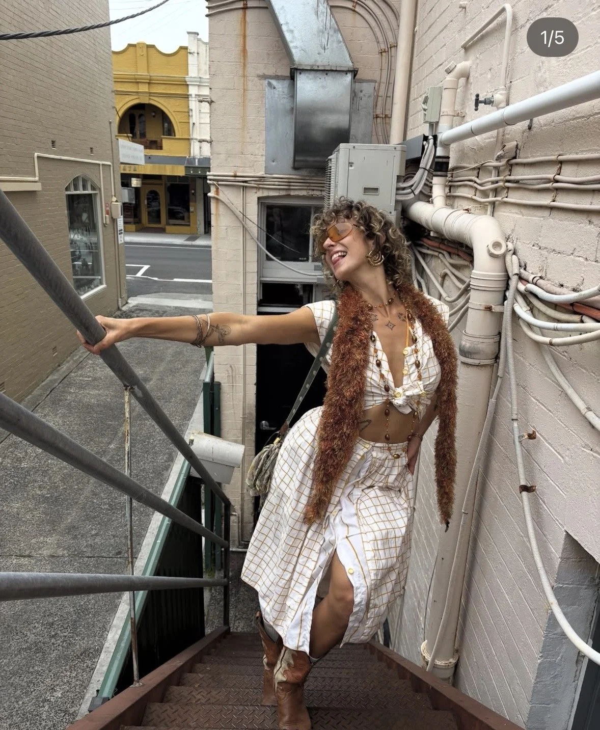 A woman with curly hair, wearing sunglasses, a patterned crop top, and a flowy skirt, standing on an outdoor staircase near a wall with exposed pipes, smiling and posing.