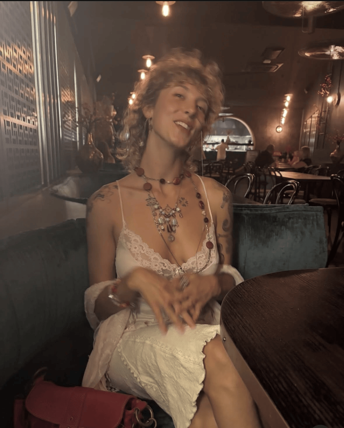 A woman with curly blonde hair, multiple necklaces, tattoos on her arms, wearing a white lace top, sitting at a table in a dimly lit restaurant or cafe.