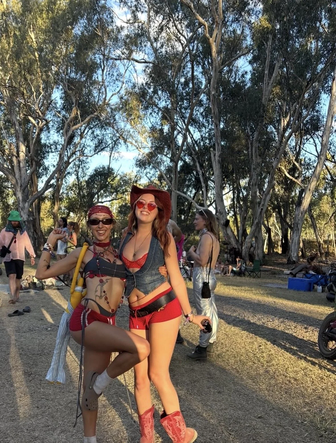 Two women dressed in festival outfits, smiling with arms around each other, standing outdoors in a wooded area during the daytime, surrounded by other festival-goers.