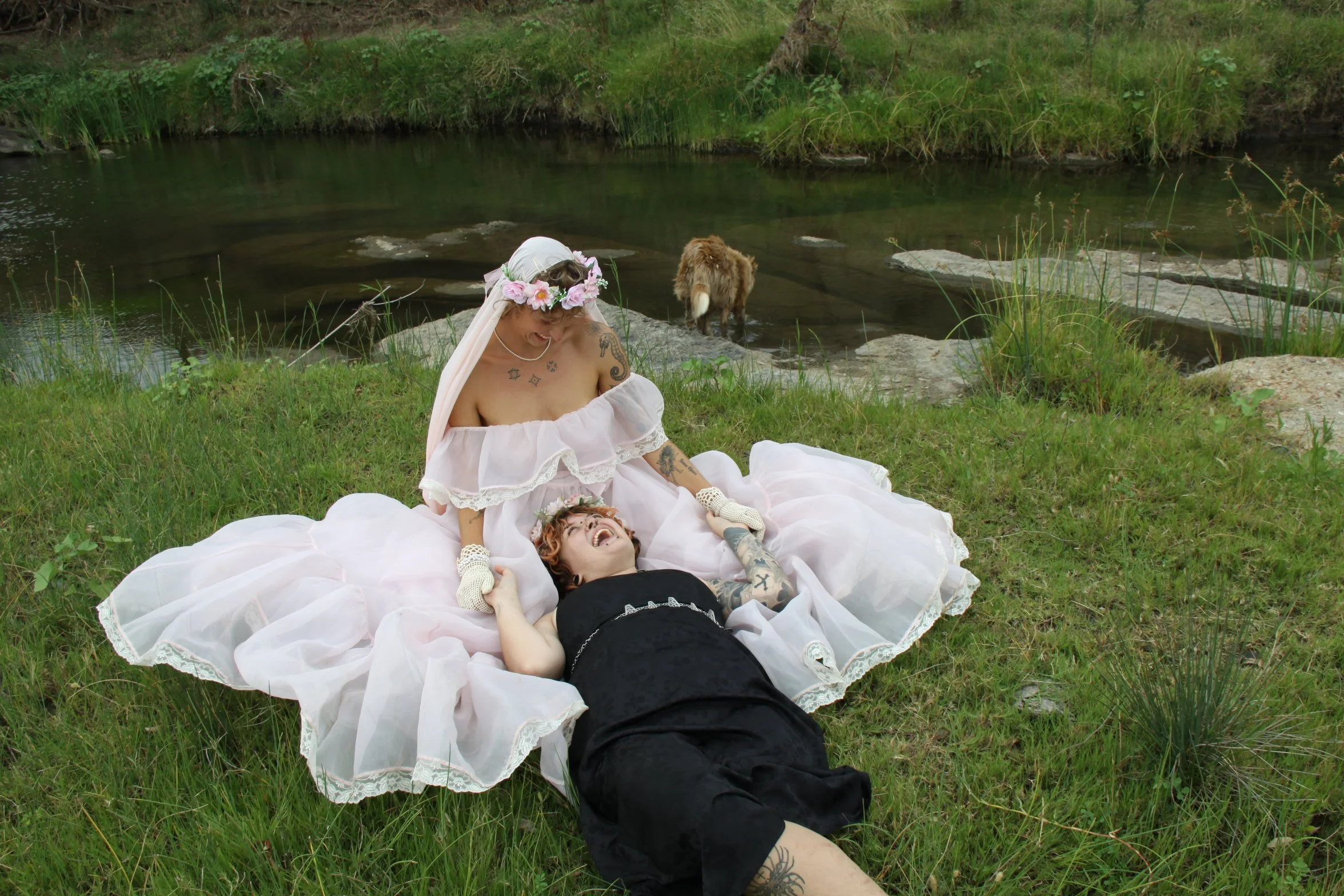 People dressed in whimsical, fairy-tale costumes lying on the grass near a river, smiling and laughing, with a dog in the water nearby and lush greenery surrounding them.