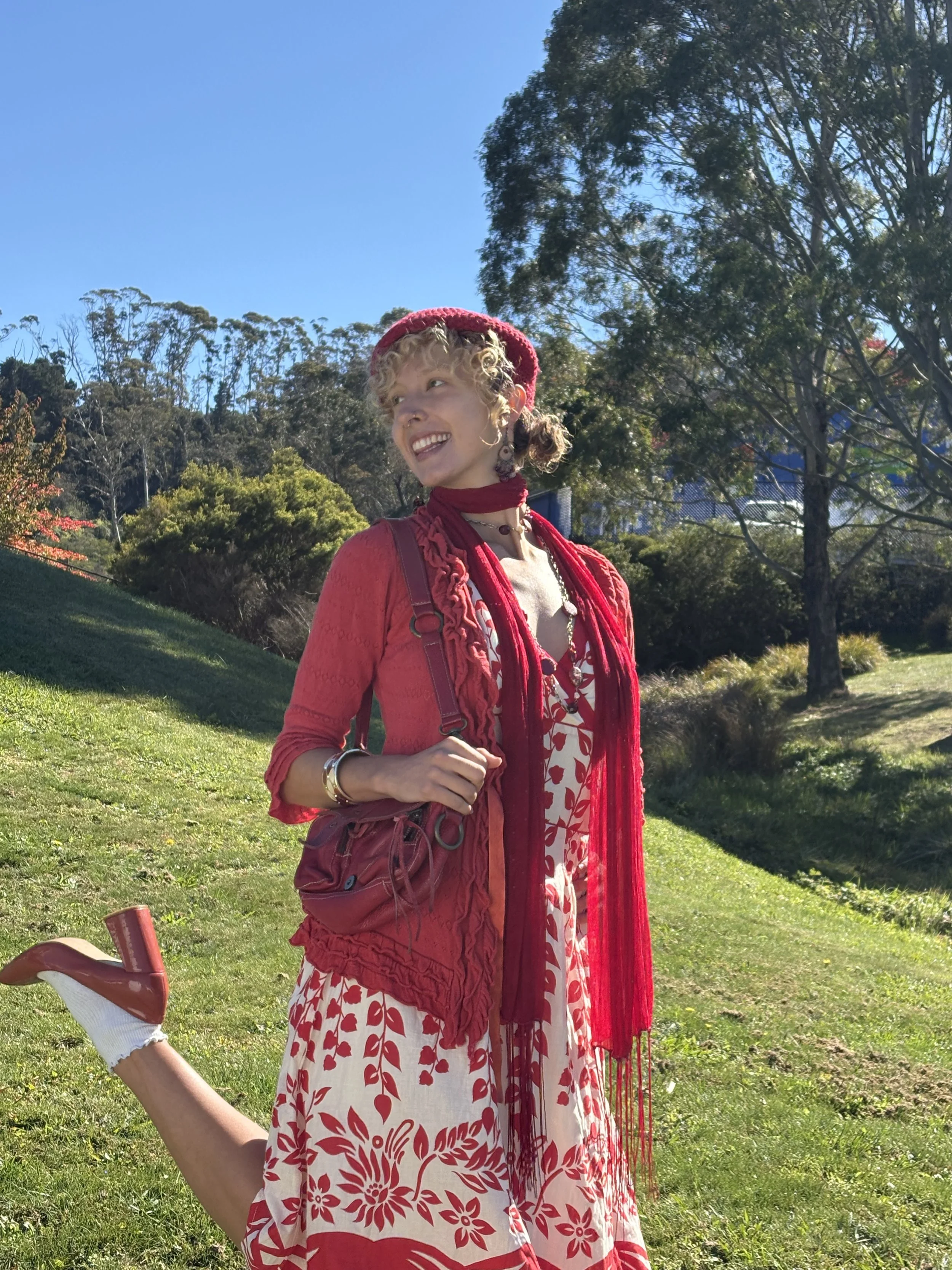 A woman with curly blonde hair wearing a red hat, red scarf, and a dress with a red floral pattern standing outdoors on green grass with trees and a blue sky in the background. She appears to be smiling and enjoying a sunny day.