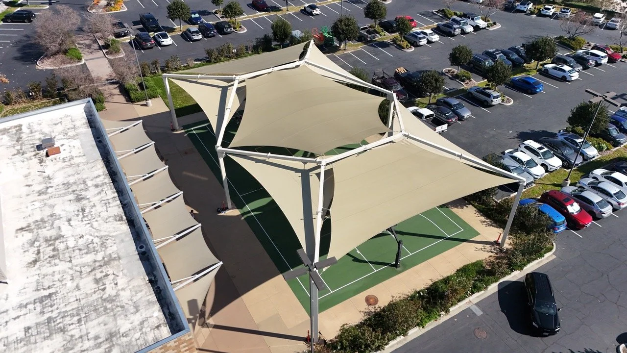 An aerial view of a parking lot with a shaded basketball court and a building with a white roof.