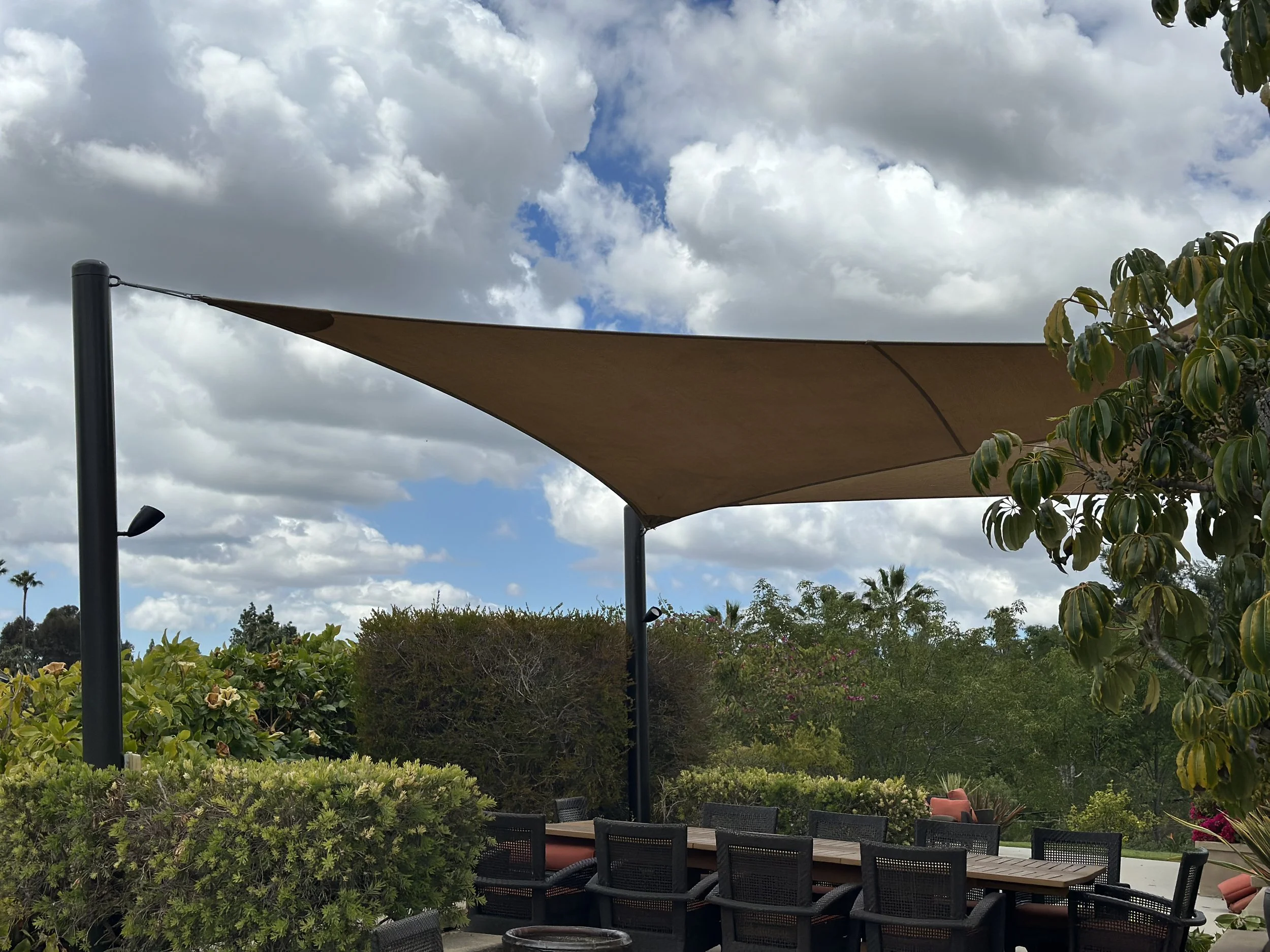 Outdoor patio with a canopy shade, surrounded by green bushes and trees under a partly cloudy sky.
