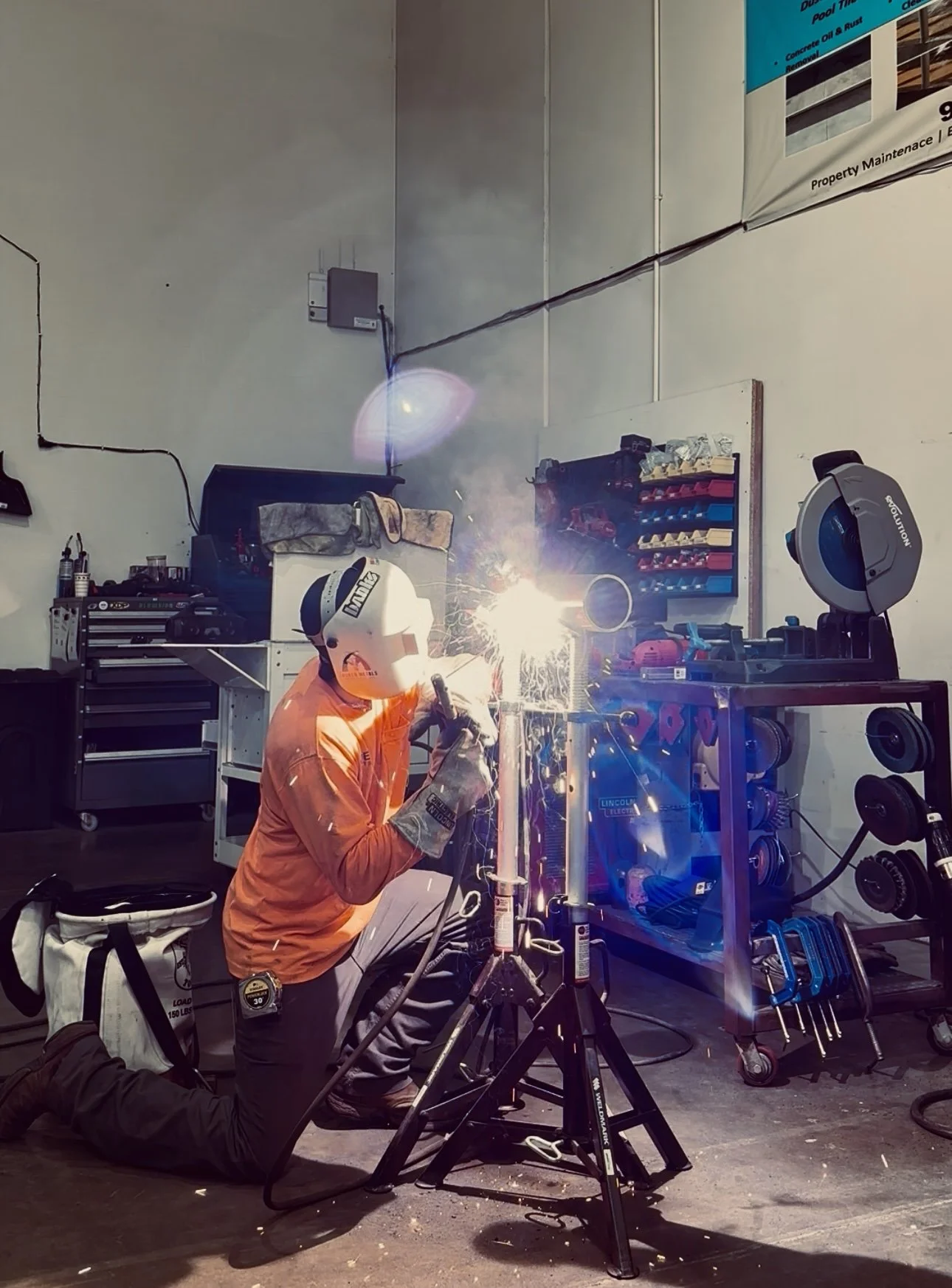 Person welding metal pipe in a workshop, wearing safety gear including helmet, gloves, and protective clothing, with sparks flying around.