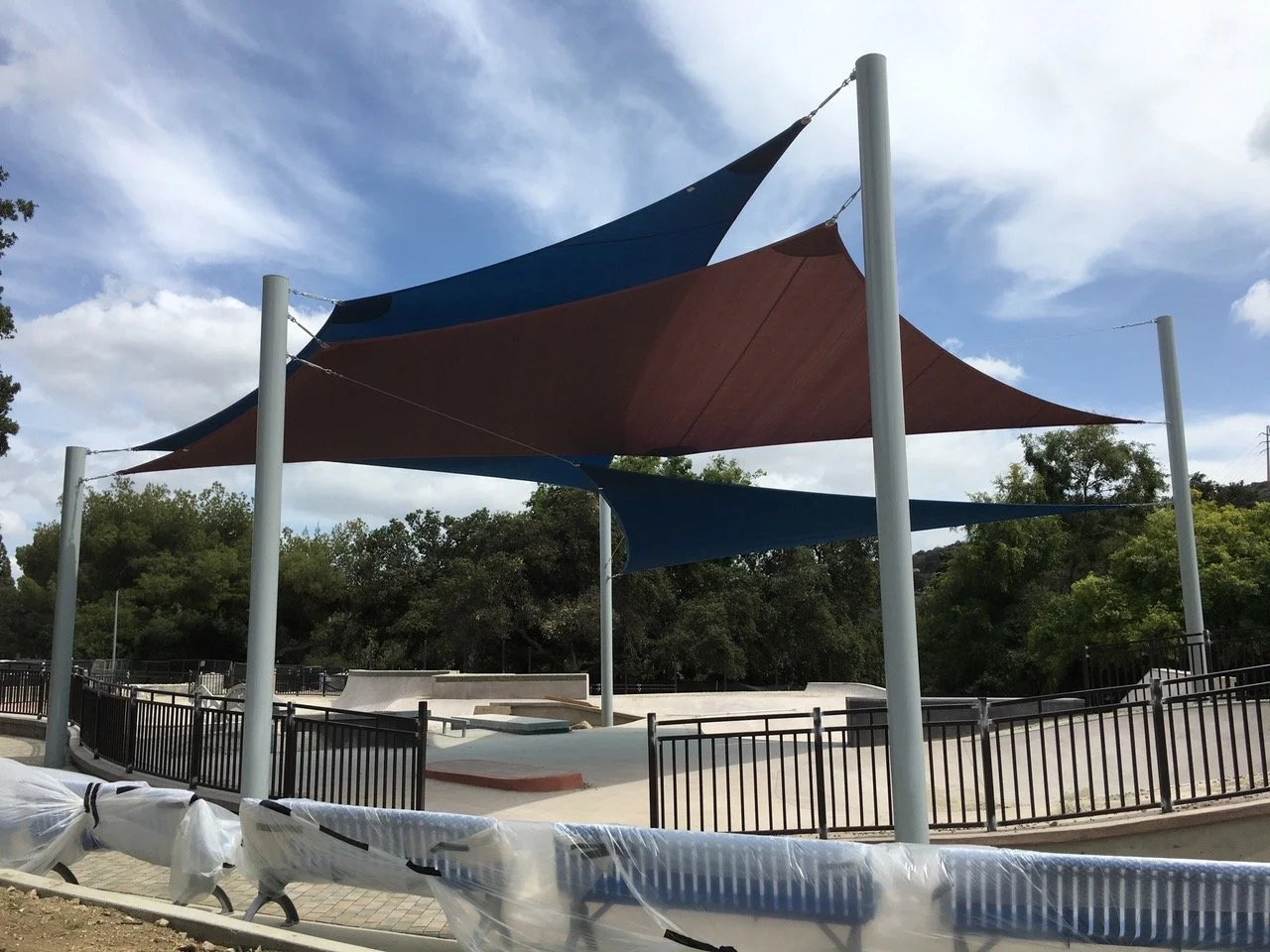 Shade structures with sail-like fabric shades over a skatepark, surrounded by trees and a black metal fence.
