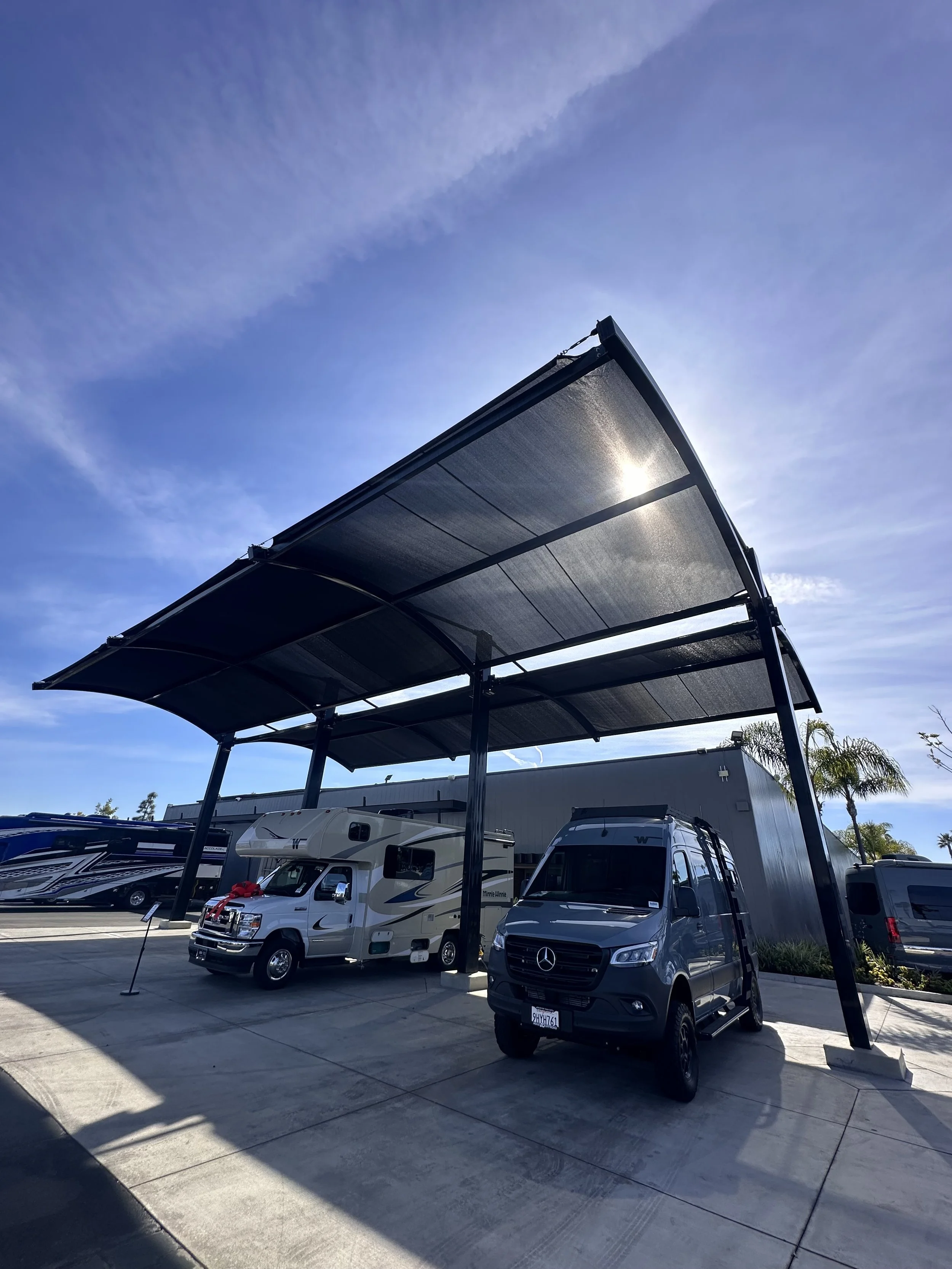 A parking lot with three recreational vehicles (RVs) parked under a large black solar panel canopy against a blue sky with wispy clouds.