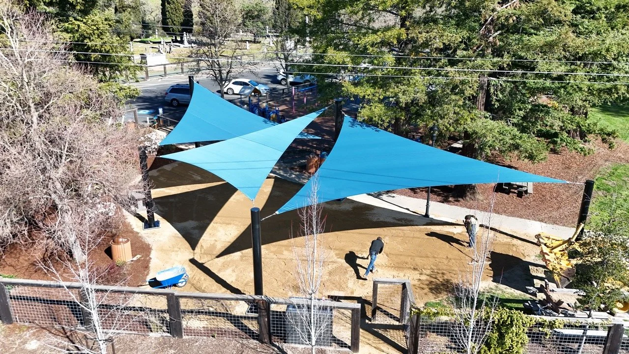 An outdoor playground area with blue shade sails overhead, dirt ground, trees, and parked cars nearby.
