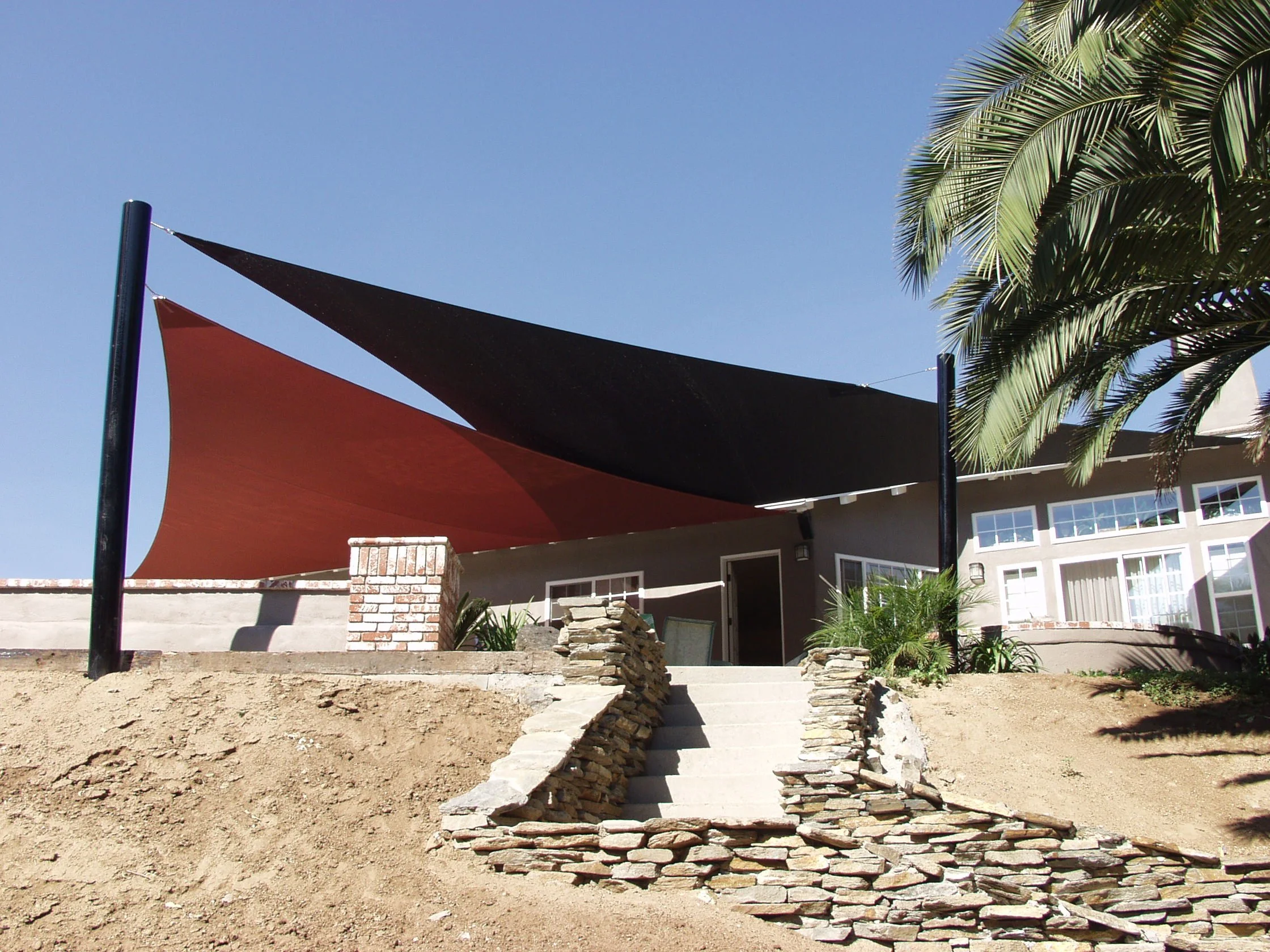A house with a backyard patio featuring two overlapping sun shades, one black and one red, attached to poles. Stone steps lead up to the patio, with a sand and rock landscape surrounding the stairs. A large palm tree is on the right side of the image, casting shadows.