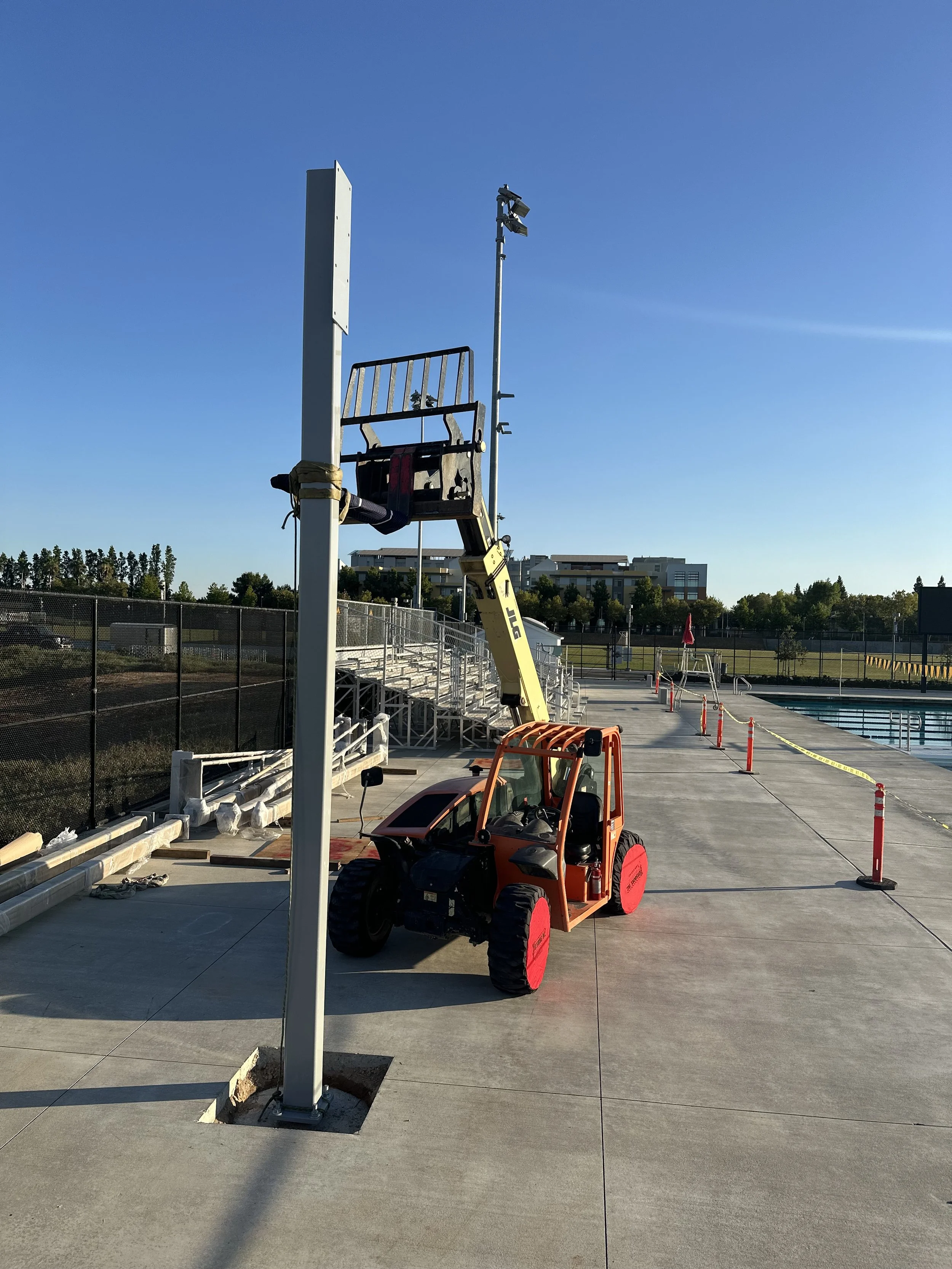 A forklift lifting a large white billboard post beside an outdoor swimming pool with surrounding decking and safety cones. Blue sky and distant buildings in background.