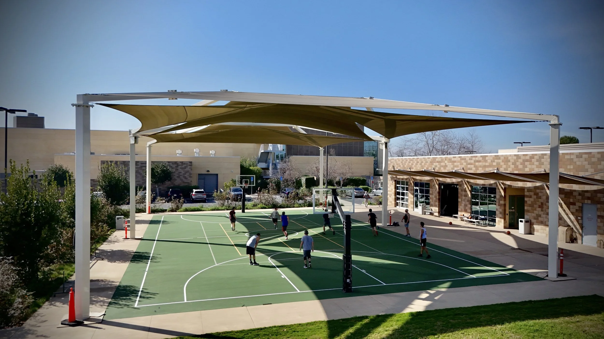 People playing basketball on an outdoor court with a shade structure and a building in the background.
