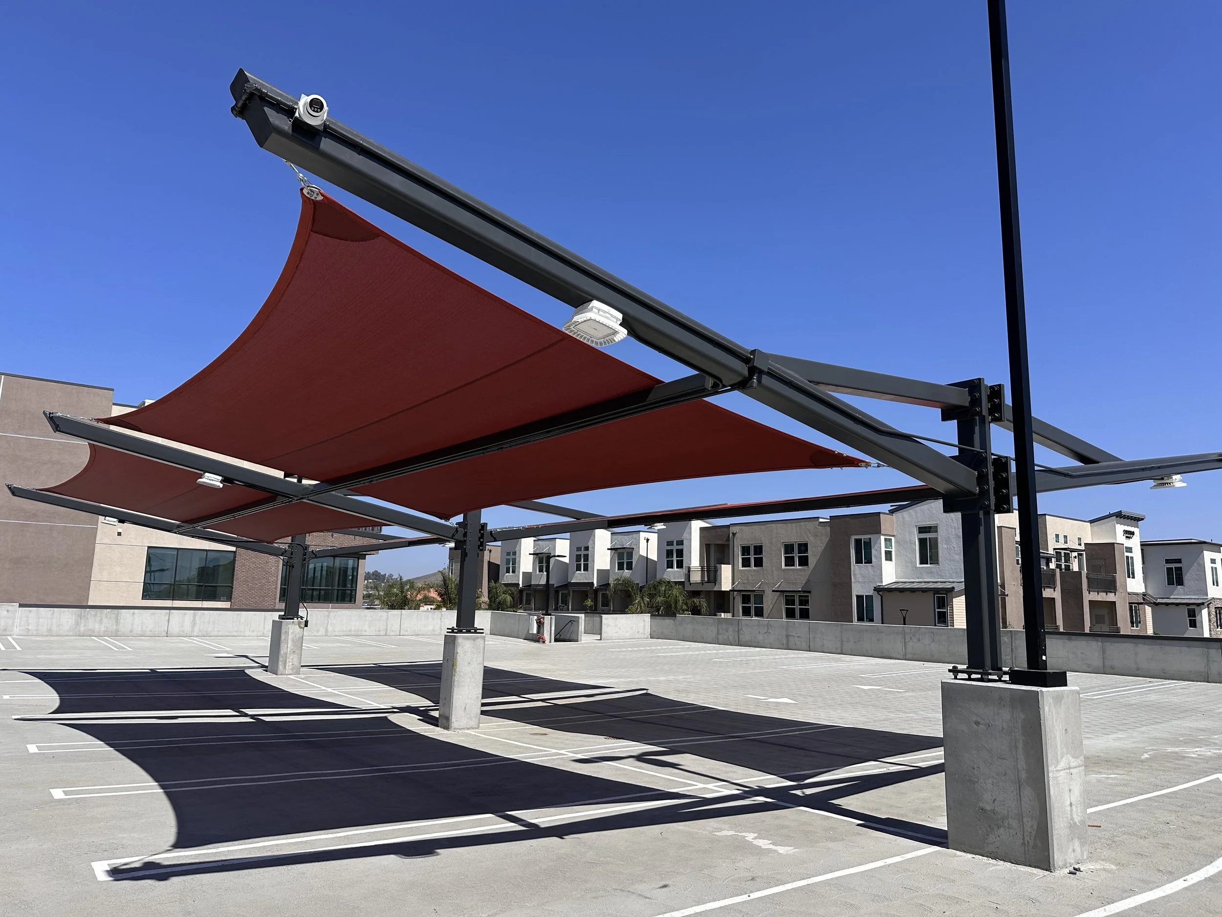 Empty parking lot with red retractable awnings overhead, residential buildings in the background, and a clear blue sky.
