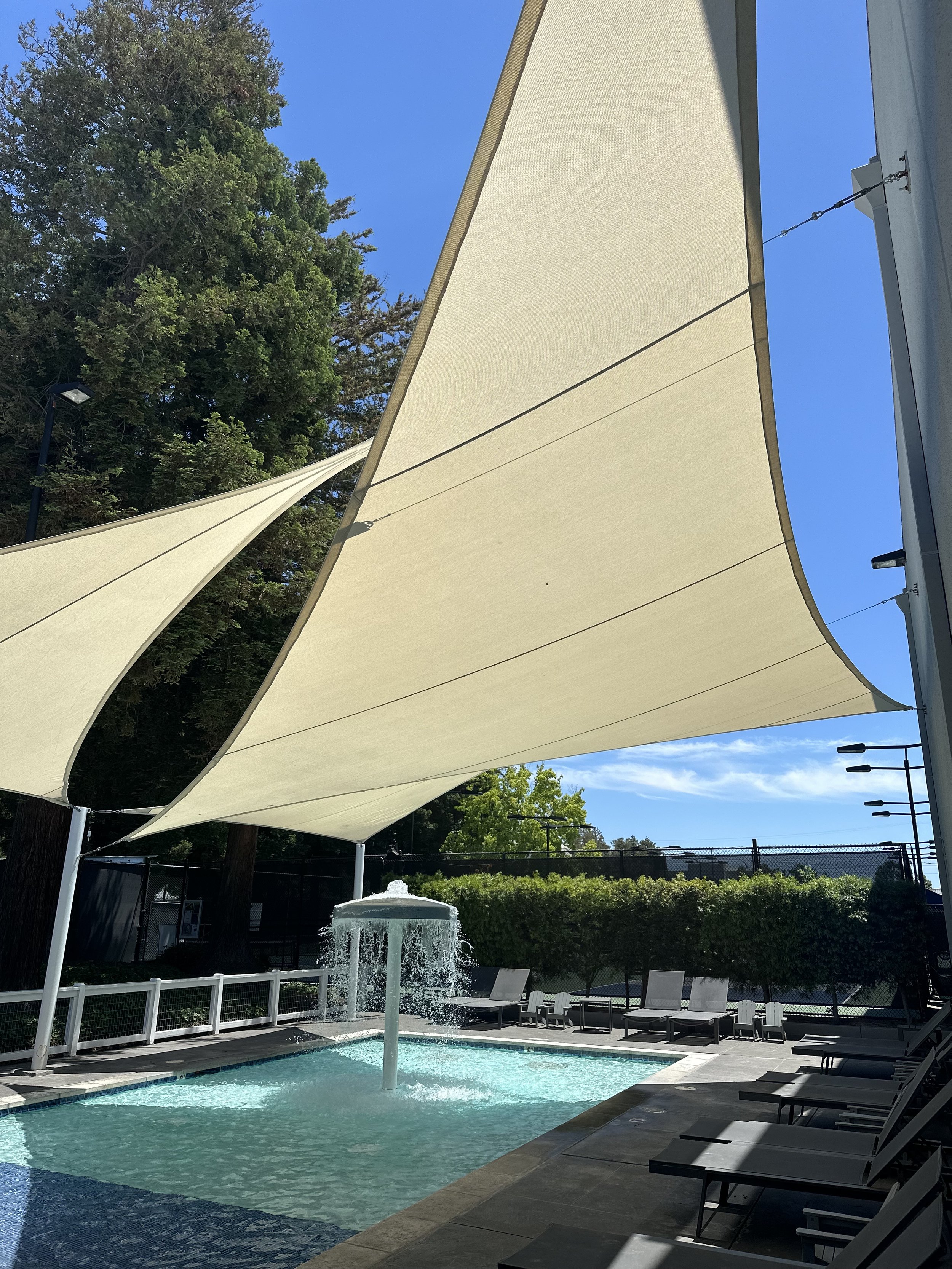 An outdoor swimming pool area with a fountain, surrounded by lounge chairs and large beige shade sails overhead on a sunny day.