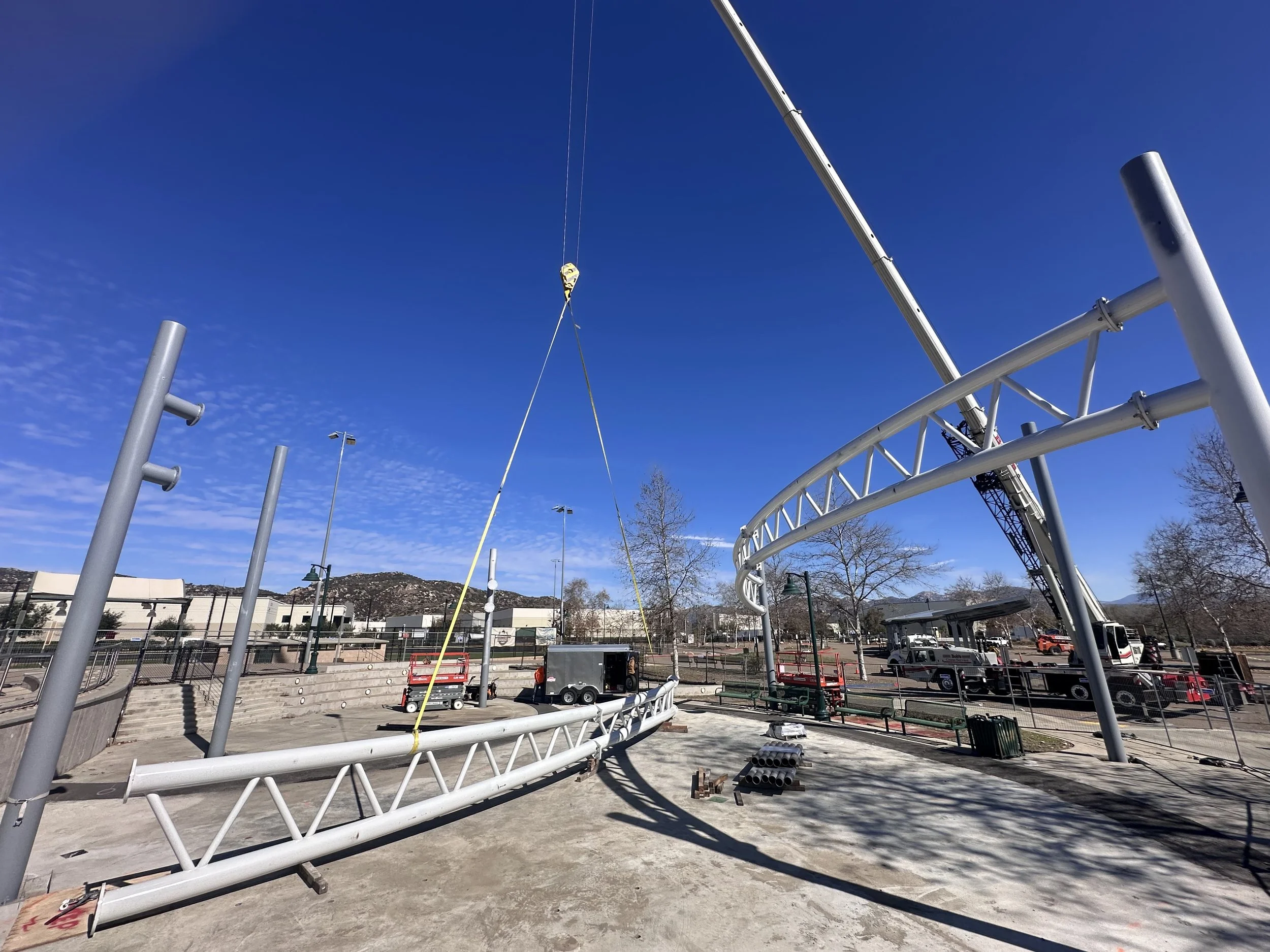 Construction site with a crane lifting a metal structure, blue sky, and some trees and trucks in the background.