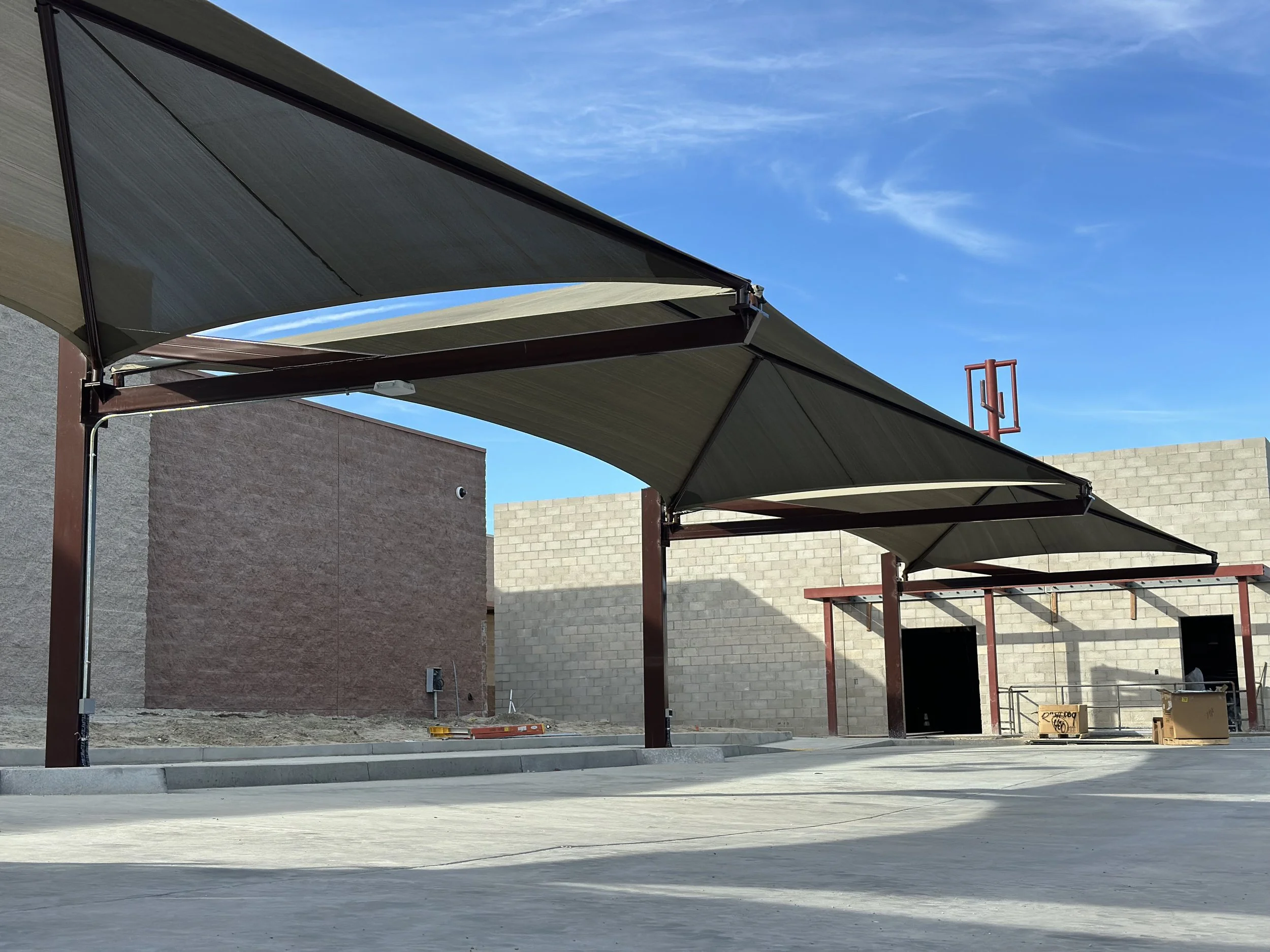 Construction site with shaded awnings, brick commercial buildings, concrete ground, and construction materials under a blue sky.