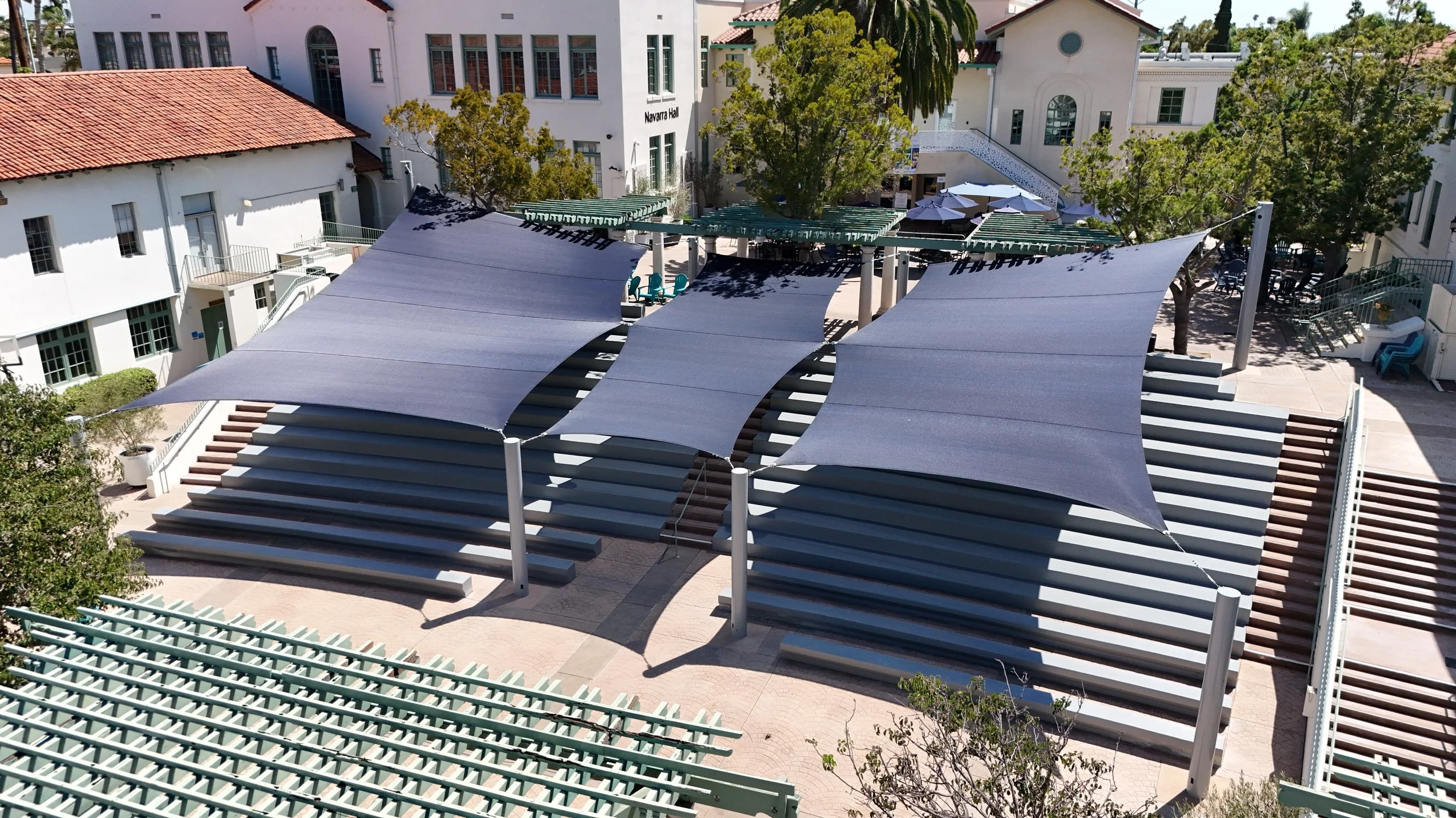 An aerial view of an outdoor amphitheater with large curved shade sails, tiered seating, and trees surrounding the area.