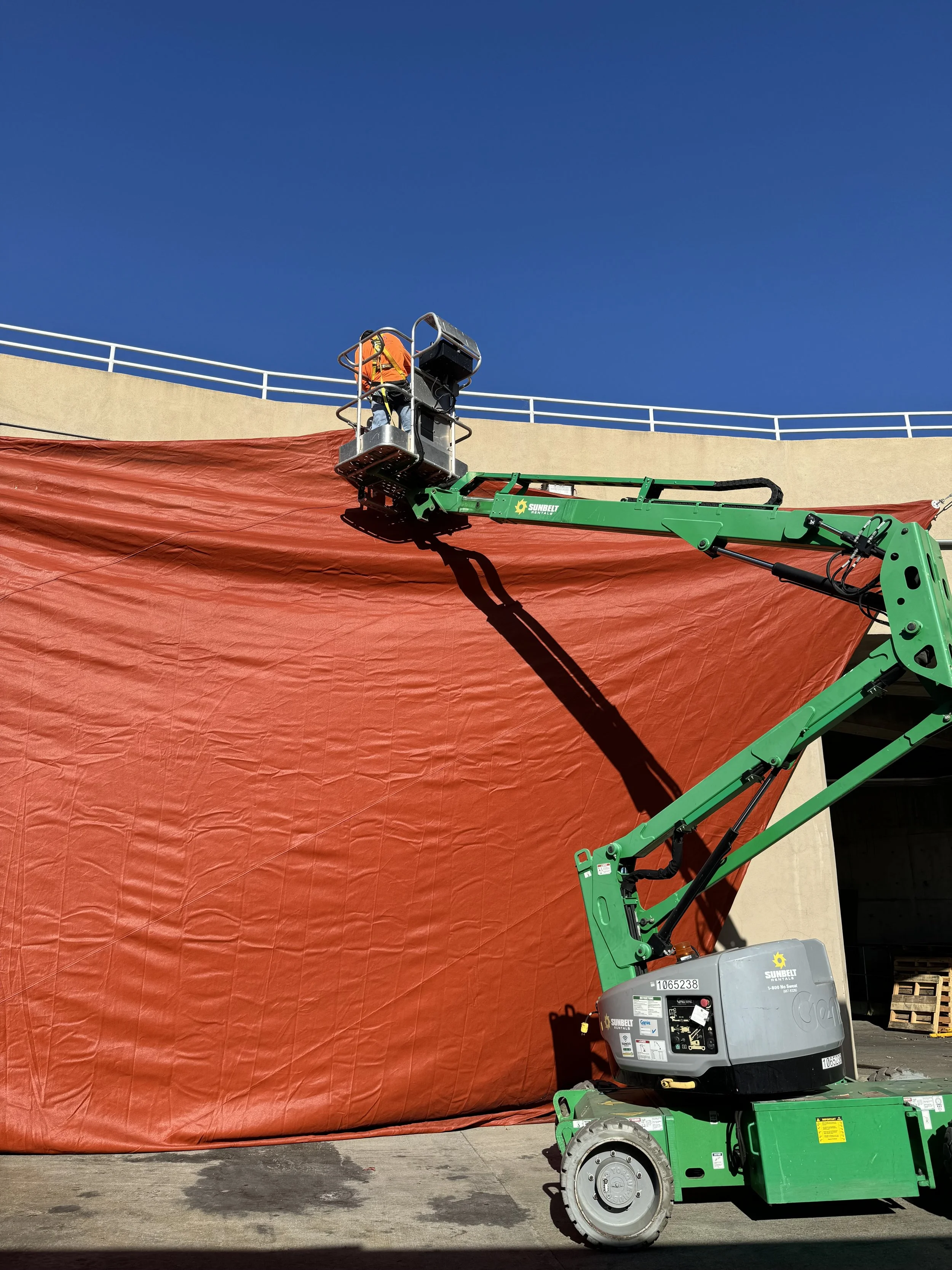 A worker operating a boom lift to hang a large orange tarp on a building exterior against a clear blue sky.